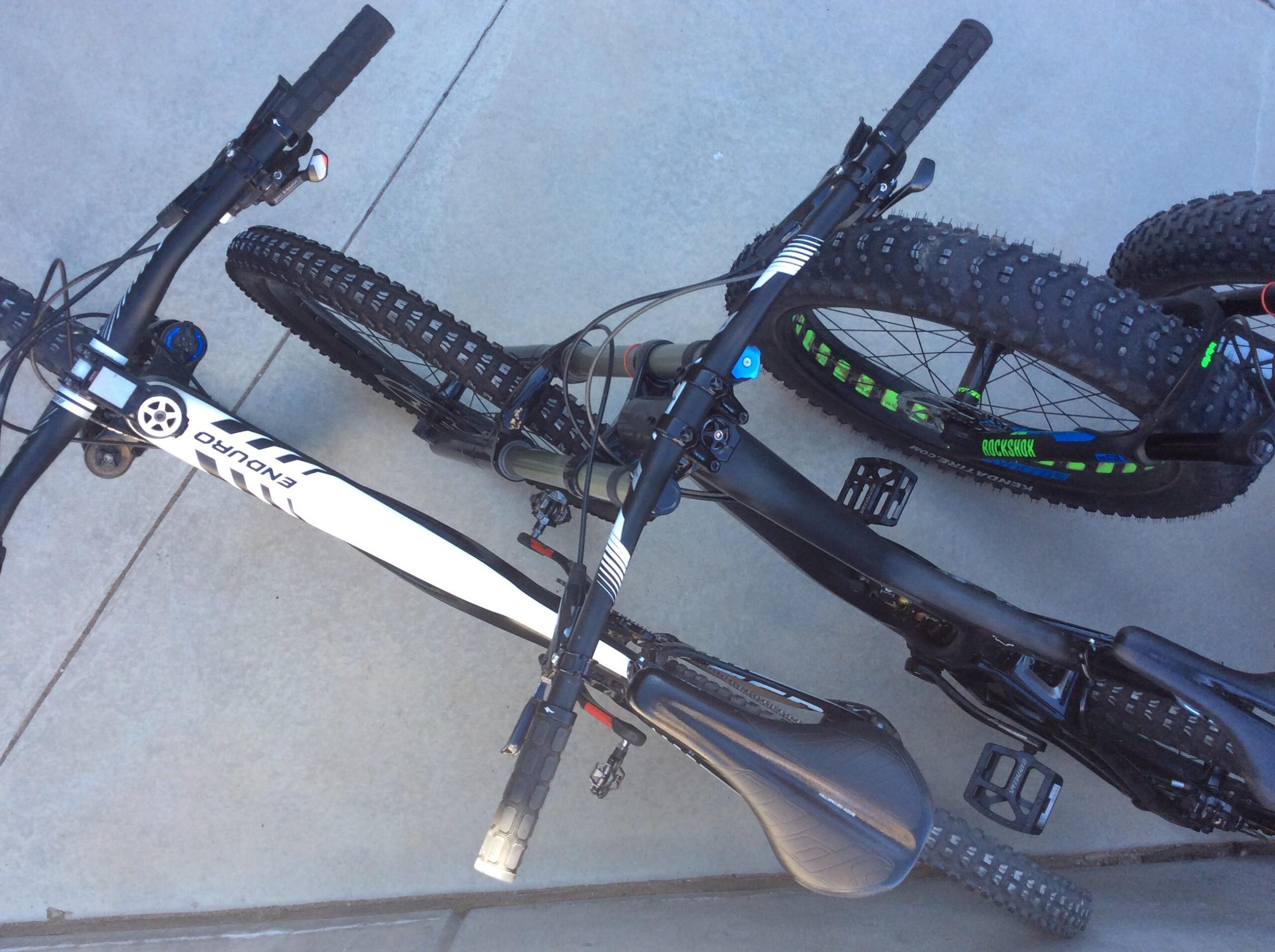Two bicycles are parked next to each other on a concrete surface. One bike has a white frame with black accents and a distinctive logo, while the other has a sleek black frame with green detailing on the tires. Both bikes feature thick, textured tires suited for off-road terrain. The handlebars and pedals of each bike are visible, showcasing their designs and grip patterns.