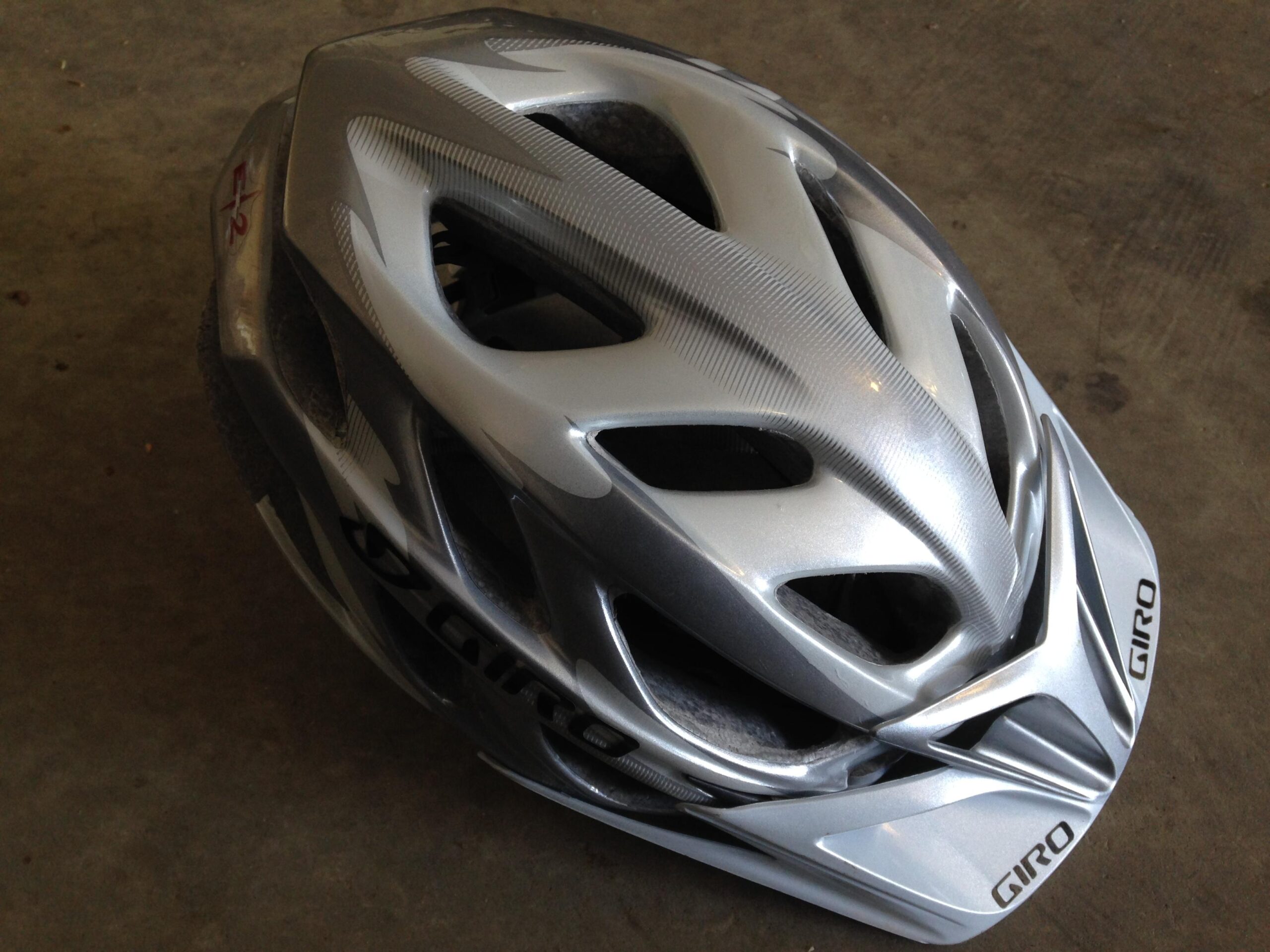 A silver Giro bicycle helmet placed on a flat surface, featuring a sleek design with ventilation openings and branding on the front. The helmet shows signs of wear, indicating previous use.