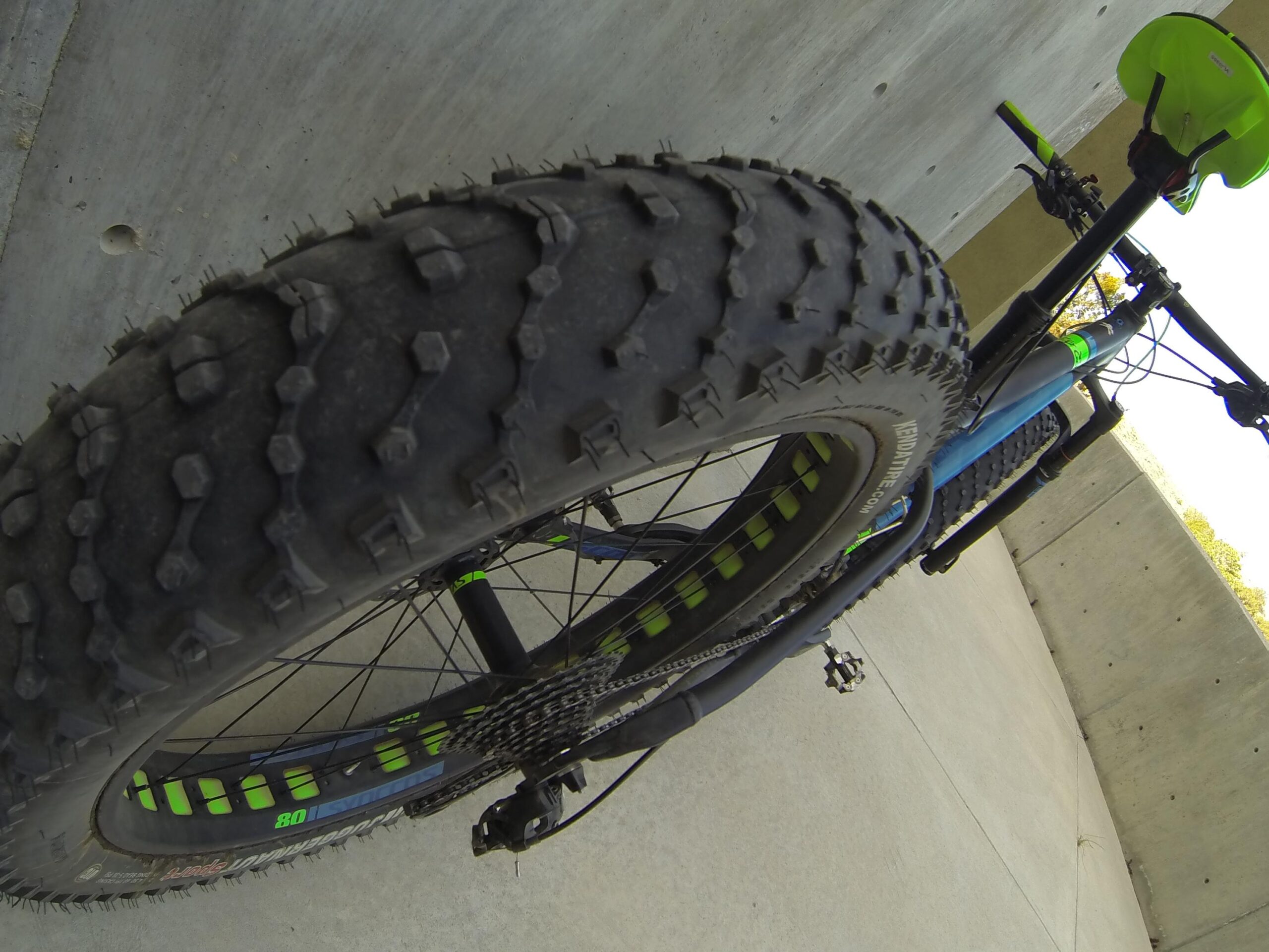 Scott Big Ed: Close-up view of a mountain bike's rear tire on a concrete surface, featuring a thick, treaded tire designed for off-road terrain, with vibrant green and blue details on the bike frame and components.