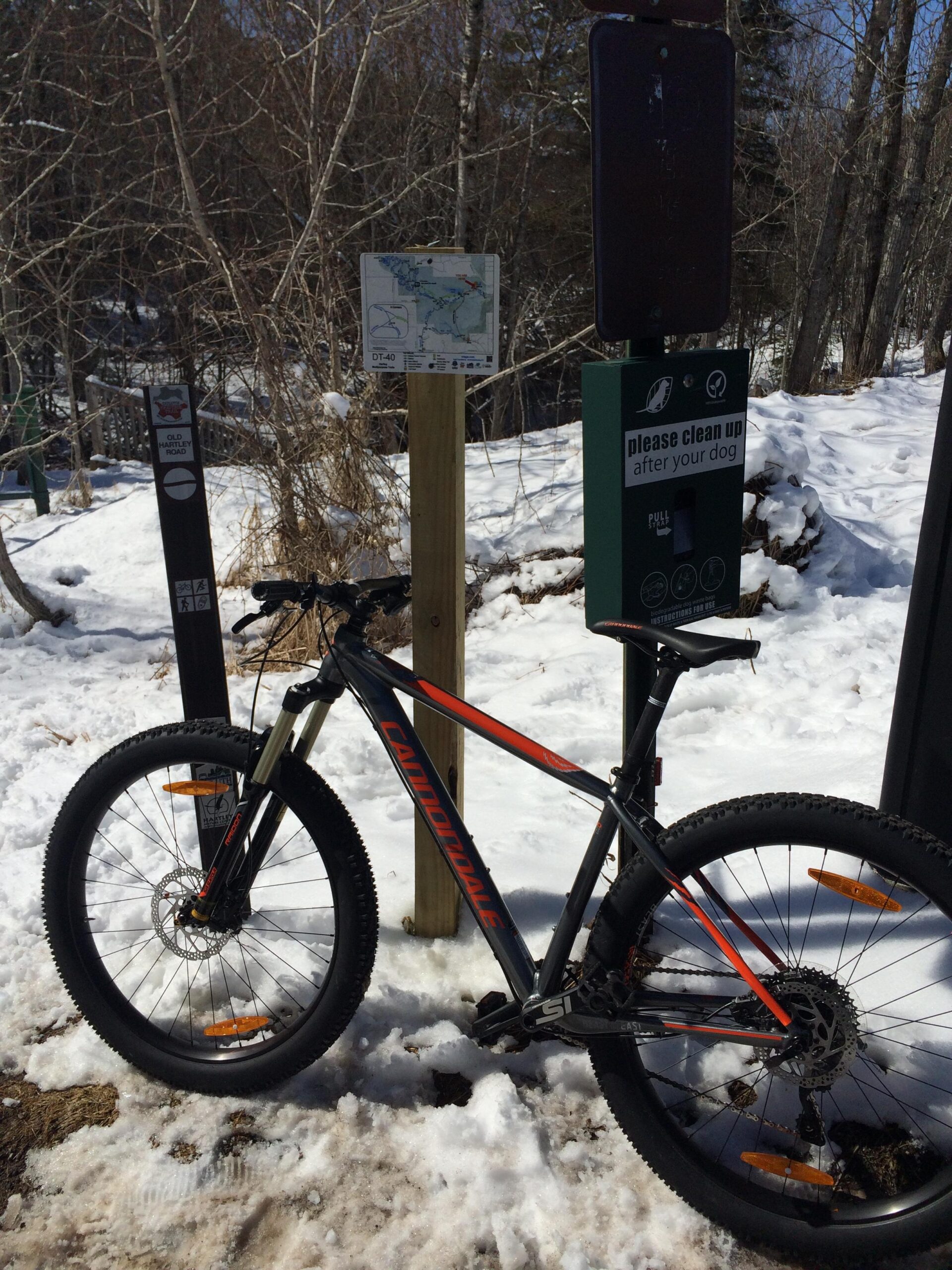A mountain bike leaning against a signpost in a snowy outdoor setting. The signpost features a map of the area labeled "DT-40" and a green dog waste station with a message reminding users to clean up after their dogs. Snow-covered ground and bare trees are visible in the background, suggesting a winter landscape. Hartley Park mountain bike trail.