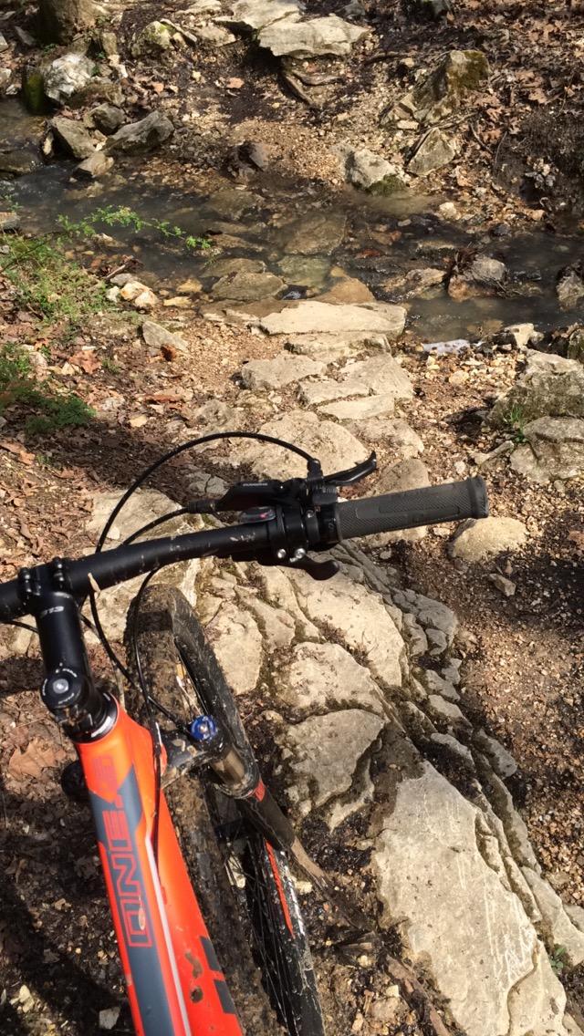 A close-up view of a mountain bike handlebar resting on a rocky path next to a small, clear stream. The surrounding area features earthy tones with scattered leaves and stones, suggesting a natural outdoor setting. Lost Valley mountain bike trail.