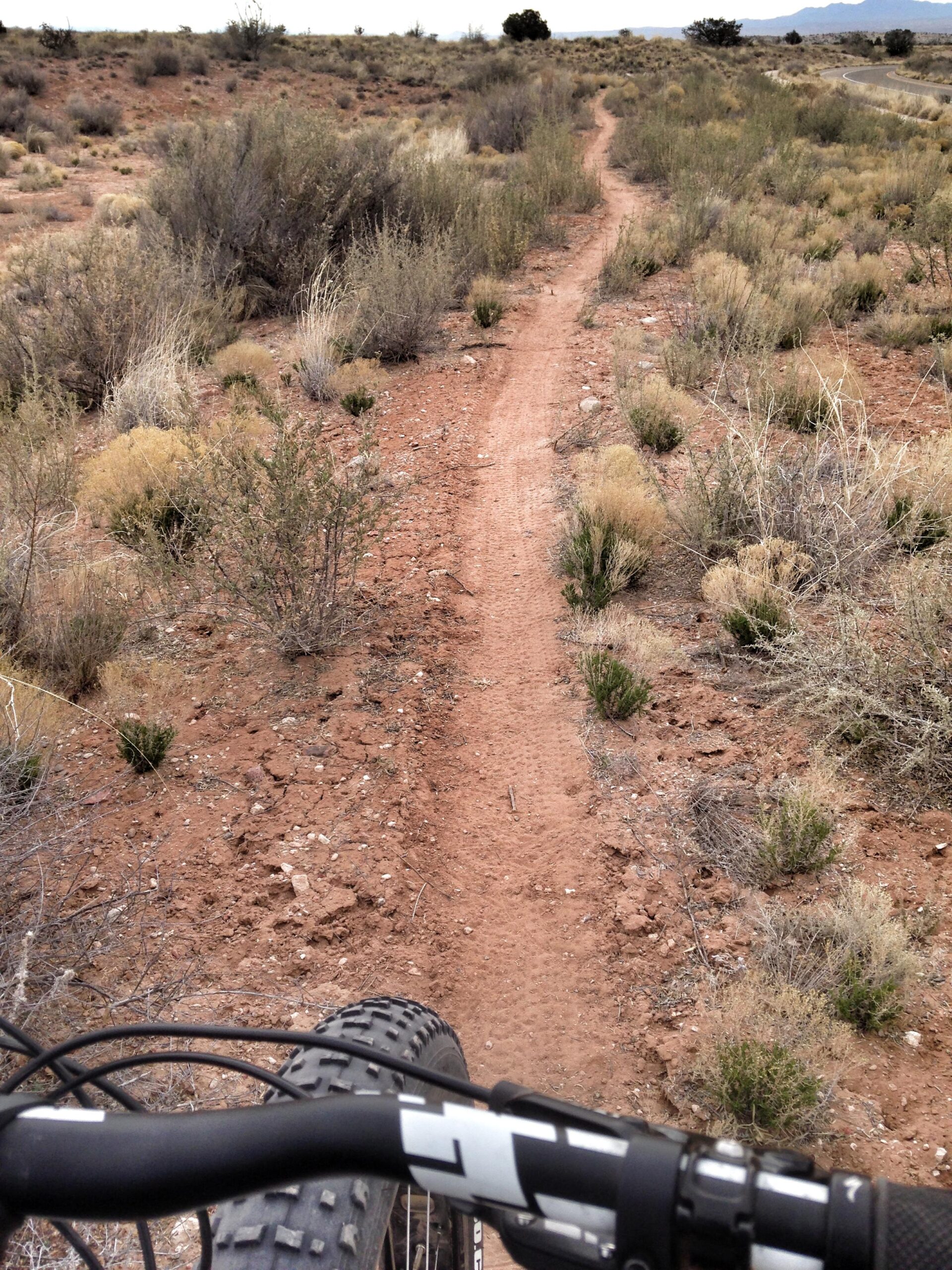 A view of a dirt biking trail winding through a desert landscape, with sparse vegetation and rugged terrain. The image shows the handlebars of a bike in the foreground, emphasizing the trail's path ahead. Parkway Fatbike trail mountain bike trail.