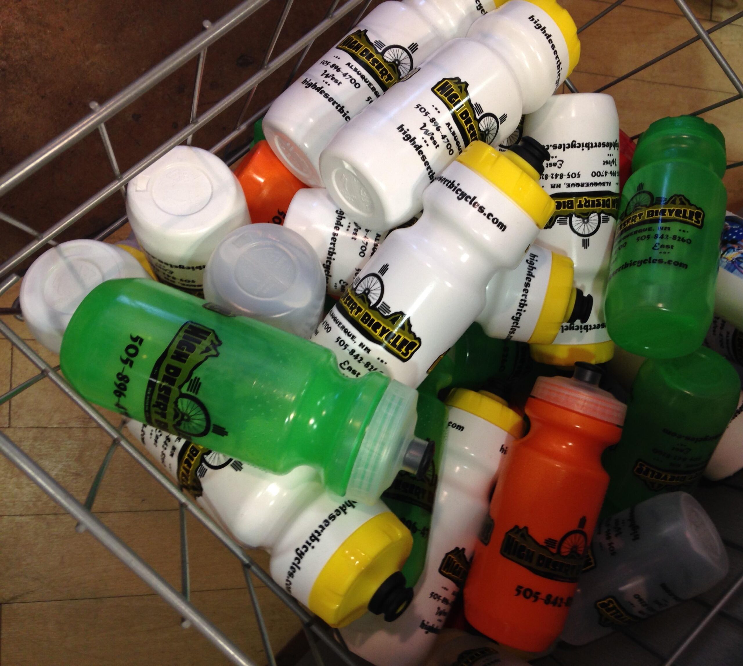 A close-up view of a wire basket filled with various plastic water bottles in different colors, including white, green, orange, and yellow. The bottles feature the logo and contact information for a business called "High Desert Bicycles." Some bottles are upright while others are partially stacked or lying on their sides.