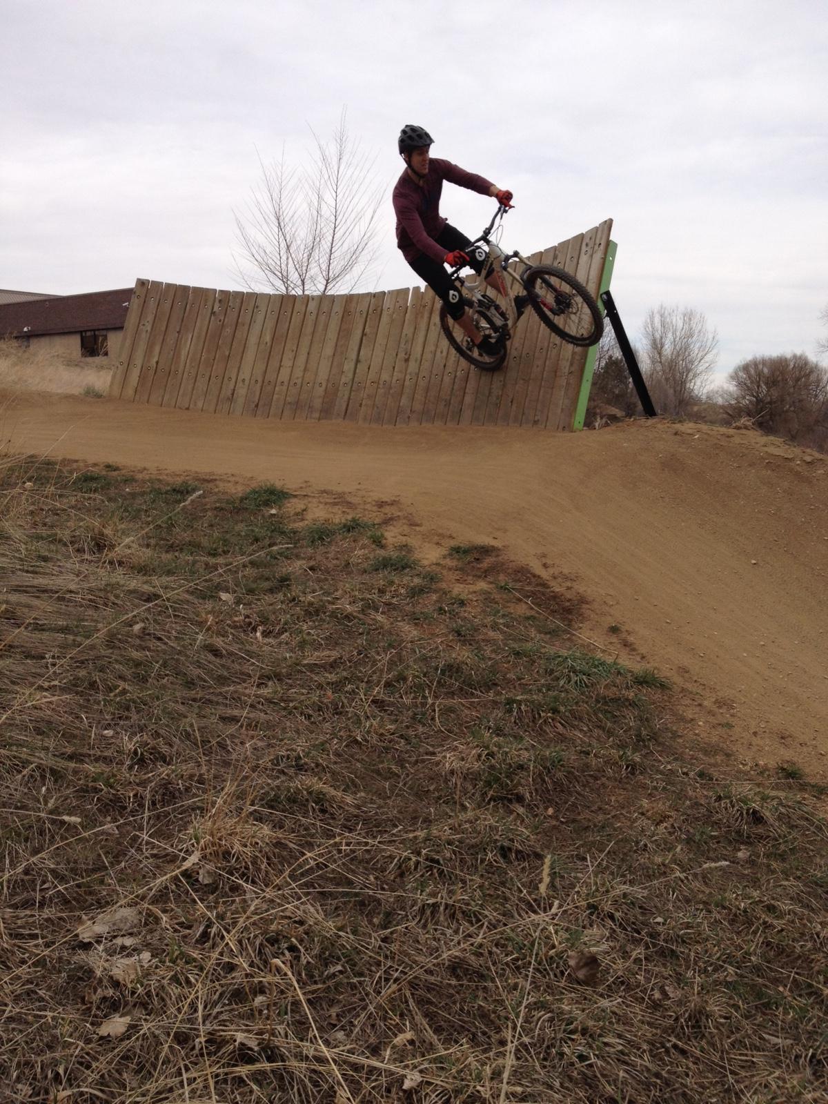 Giant Reign 2: A mountain biker performs a jump on a wooden ramp, captured mid-air against a cloudy sky. The rider is wearing a helmet and a long-sleeve shirt, with a dirt path and grass in the foreground. The wooden ramp has a curved design, leading to a downhill slope.