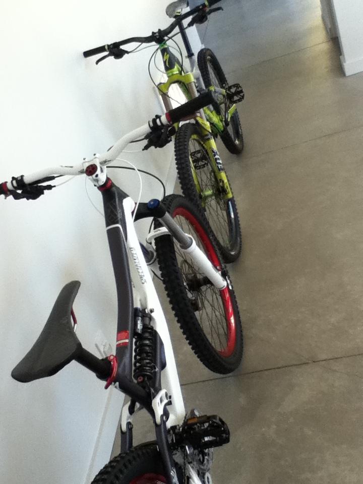 Two mountain bikes parked in a hallway, one predominantly white with red accents and the other green and black. The bikes are positioned parallel to a white wall on a concrete floor, showcasing their handlebars and tires.