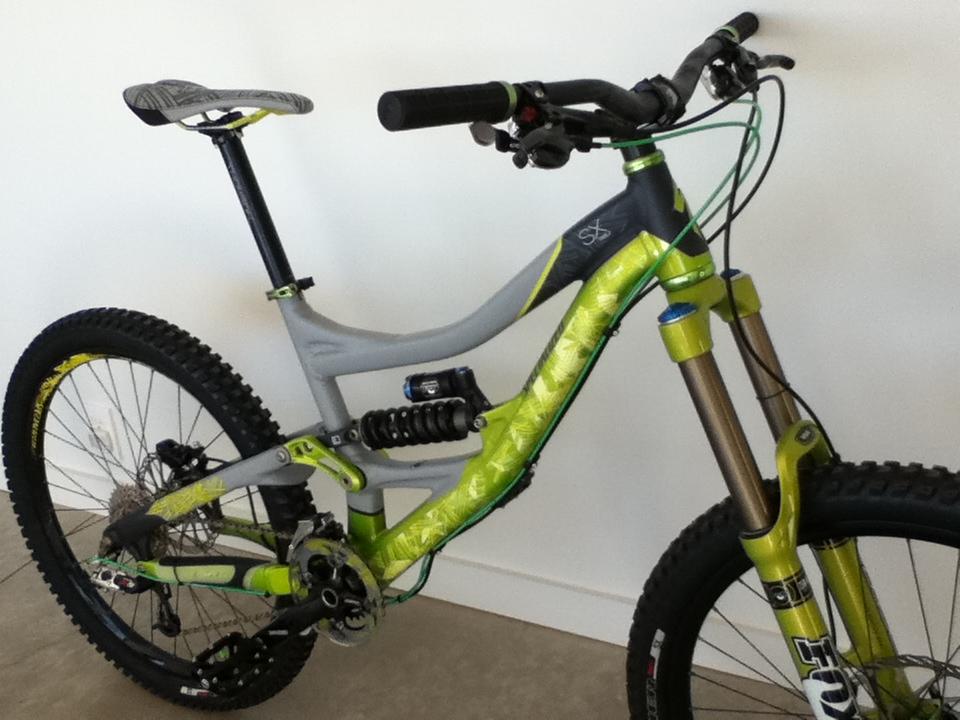 A modern mountain bike with a gray and lime green frame, featuring wide tires, a prominent shock absorber, and dual disc brakes. The bike is positioned against a plain white wall, showcasing its sleek design and components.