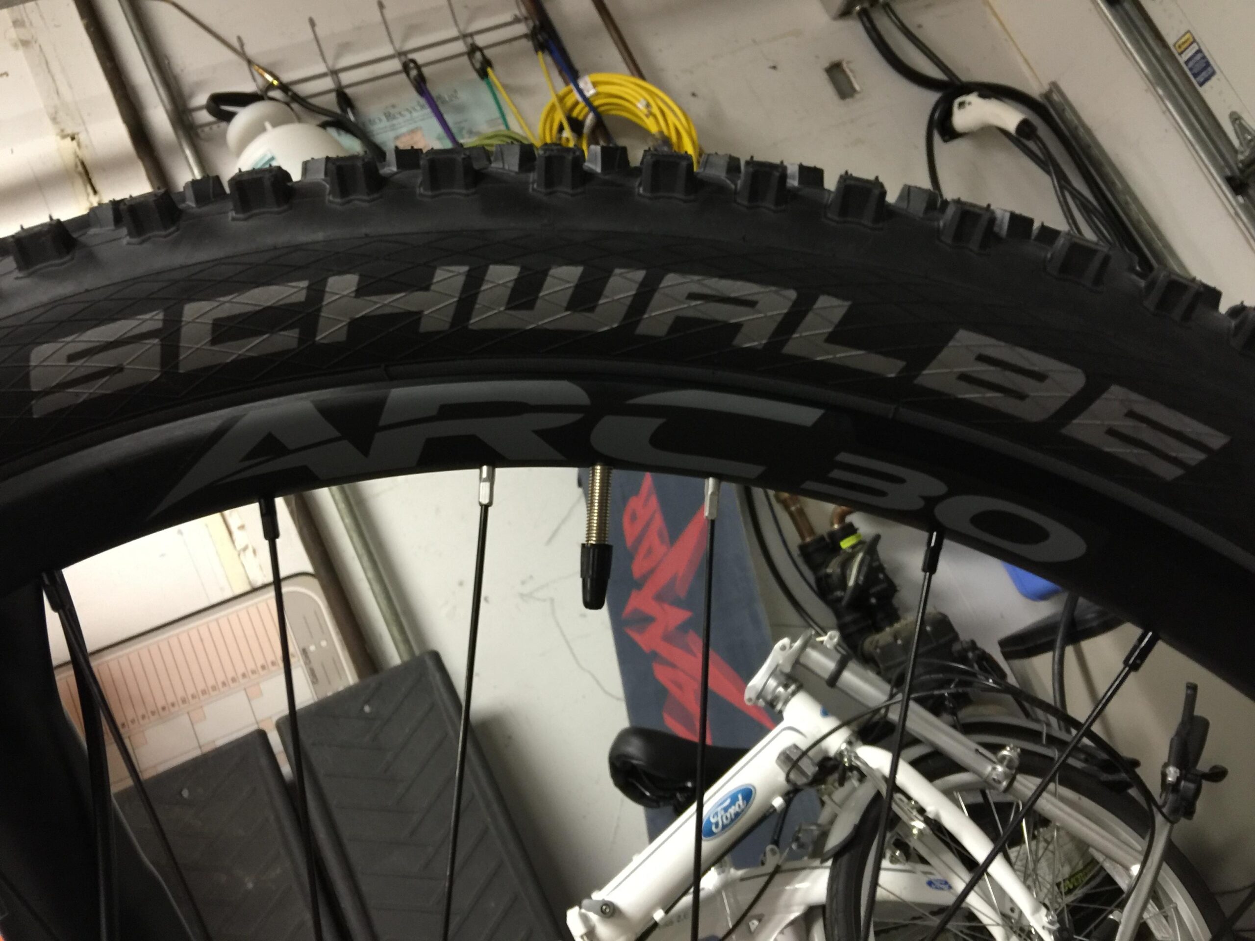 Schwalbe Hans Dampf: A close-up view of a mountain bike tire featuring the brand name "Schwalbe" and the model designation "ARC30." The tire showcases distinct tread patterns designed for off-road traction. In the background, parts of a white bicycle and garage equipment are visible.