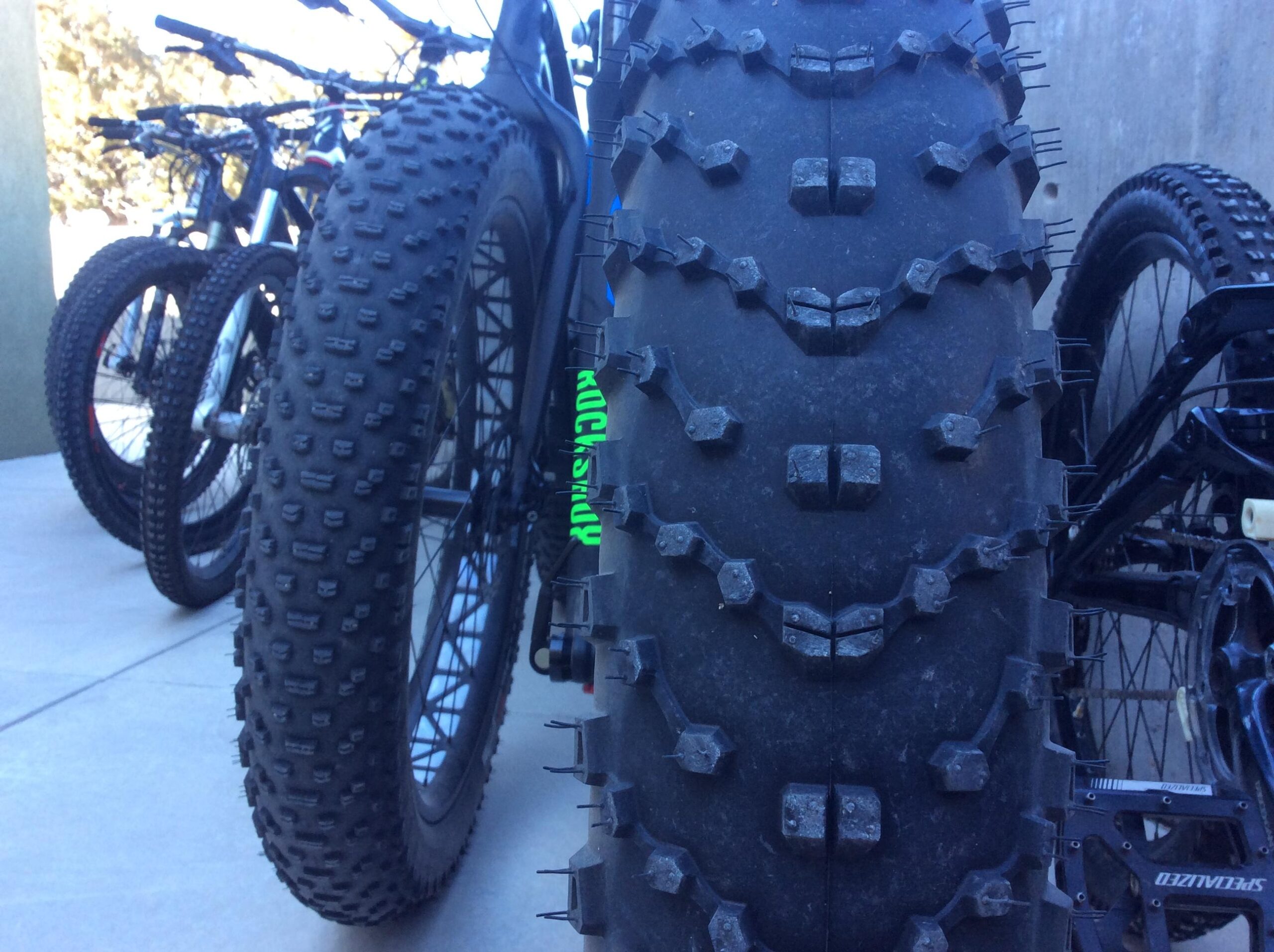 A close-up view of a large, textured bicycle tire, showcasing its tread. In the background, several other bicycles with various tire types are partially visible, all positioned on a concrete surface. The setting appears outdoors with natural lighting.
