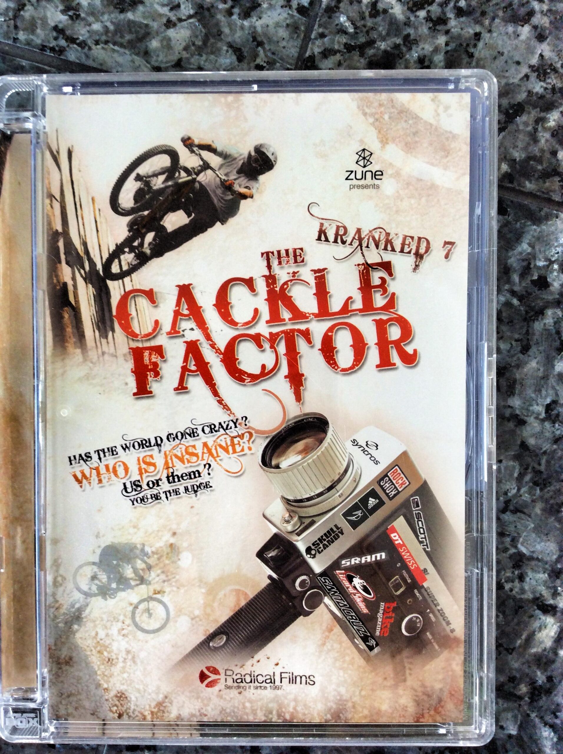 Alt text: Cover of the DVD titled "Kranked 7: The Cackle Factor," featuring an image of a mountain biker performing a trick against a backdrop of urban scenery. The design includes bold red text and phrases questioning sanity, with a camera graphic and branding elements from Zune and Radical Films.