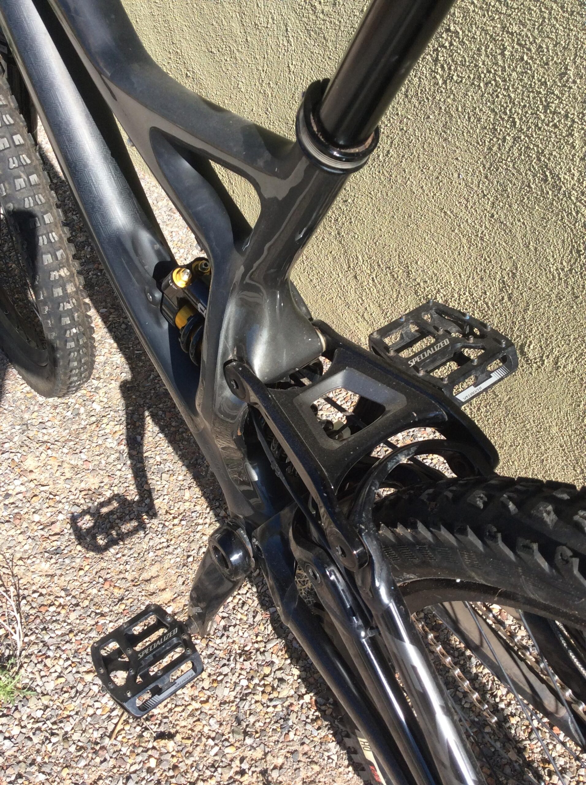 Specialized S-Works Demo 8: Close-up view of a mountain bike's pedal and frame, highlighting the black pedal, chainstay, and rear shock assembly against a textured green background. Sunlight casts a shadow on the gravel surface beneath the bike.