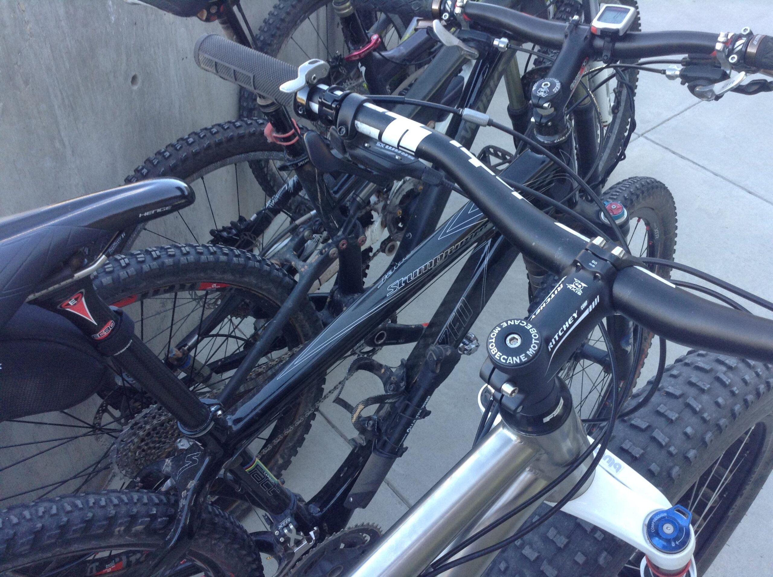 A close-up view of several mountain bikes parked together, showcasing handlebars, grips, and parts of the frames. The bikes feature thick tires suited for rugged terrain and various brands visible on the frame. The background includes a concrete wall and a paved surface.