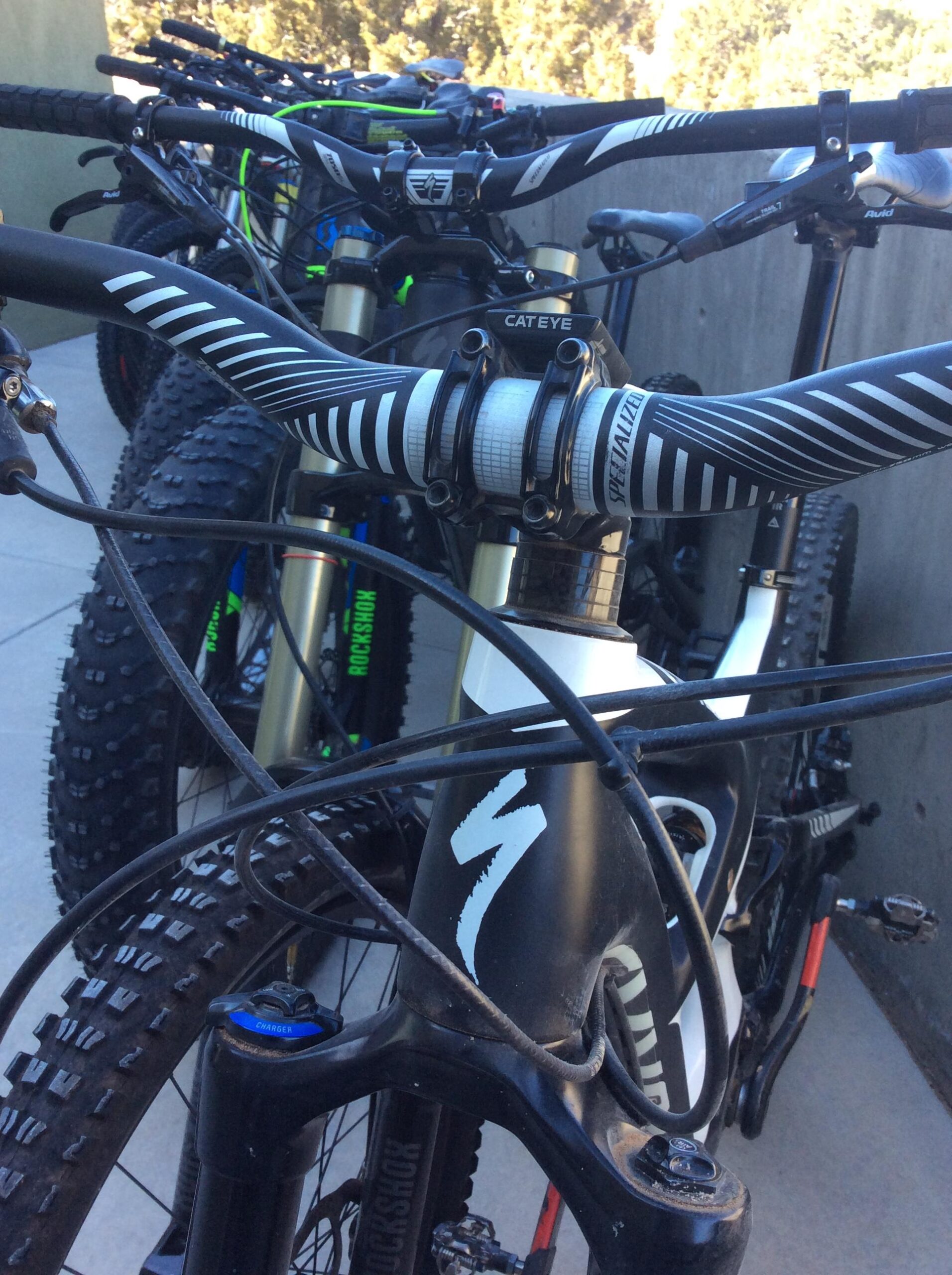 A close-up view of several mountain bikes lined up, focusing on the handlebars and front section of the nearest bike. The handlebars feature black grips with white stripes, and components are labeled with brand names such as "Specialized" and "Cateye." The scene captures the intricate details of the bike’s features, including cables and a part labeled "Charger," with a blurred background revealing more bikes.