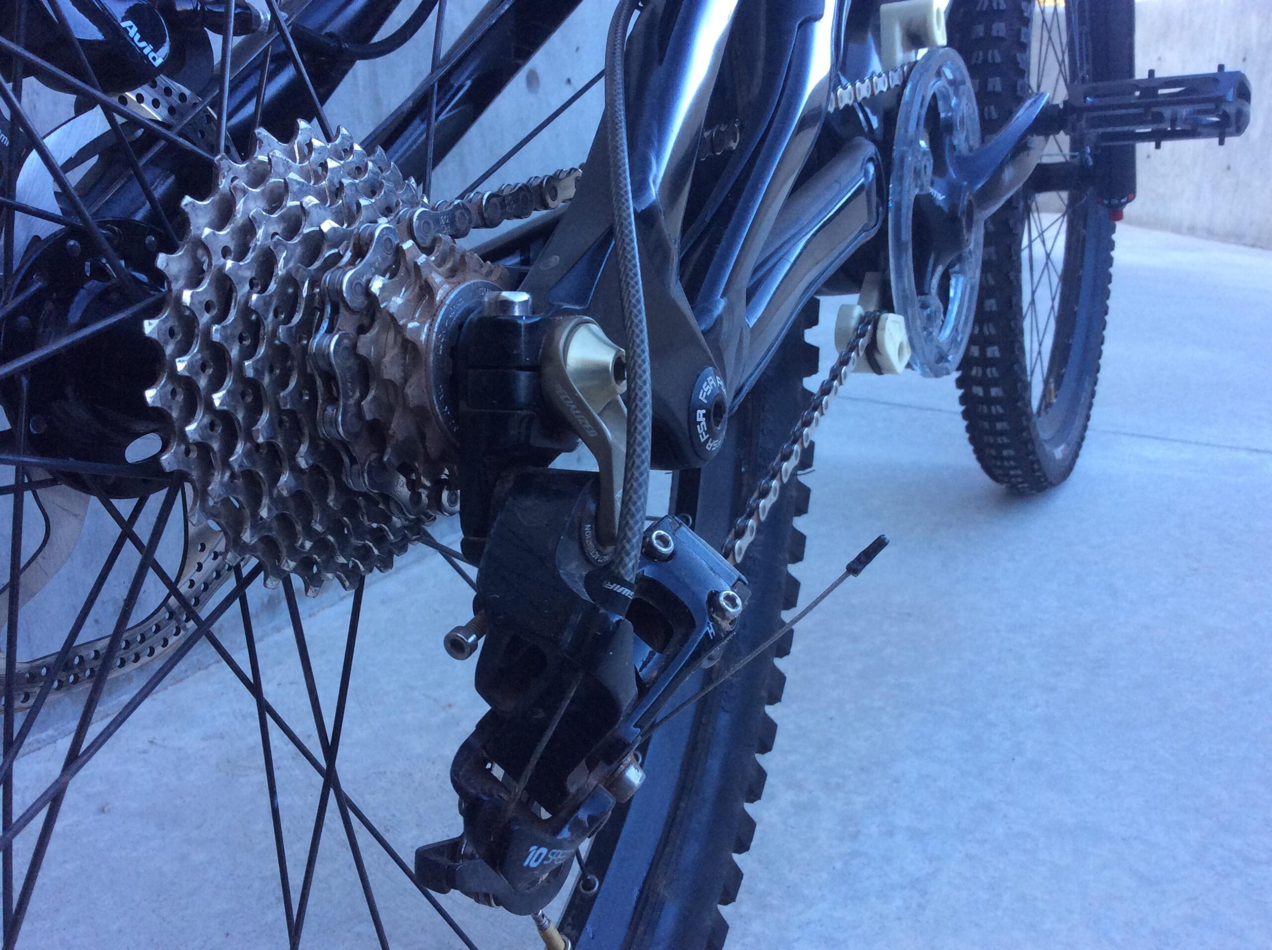Specialized S-Works Demo 8 Carbon: Close-up view of a bicycle's rear drivetrain, featuring a metallic cassette, derailleur, and chain. The image highlights the intricate details of the gear system, showing the connection between the components against a concrete background.