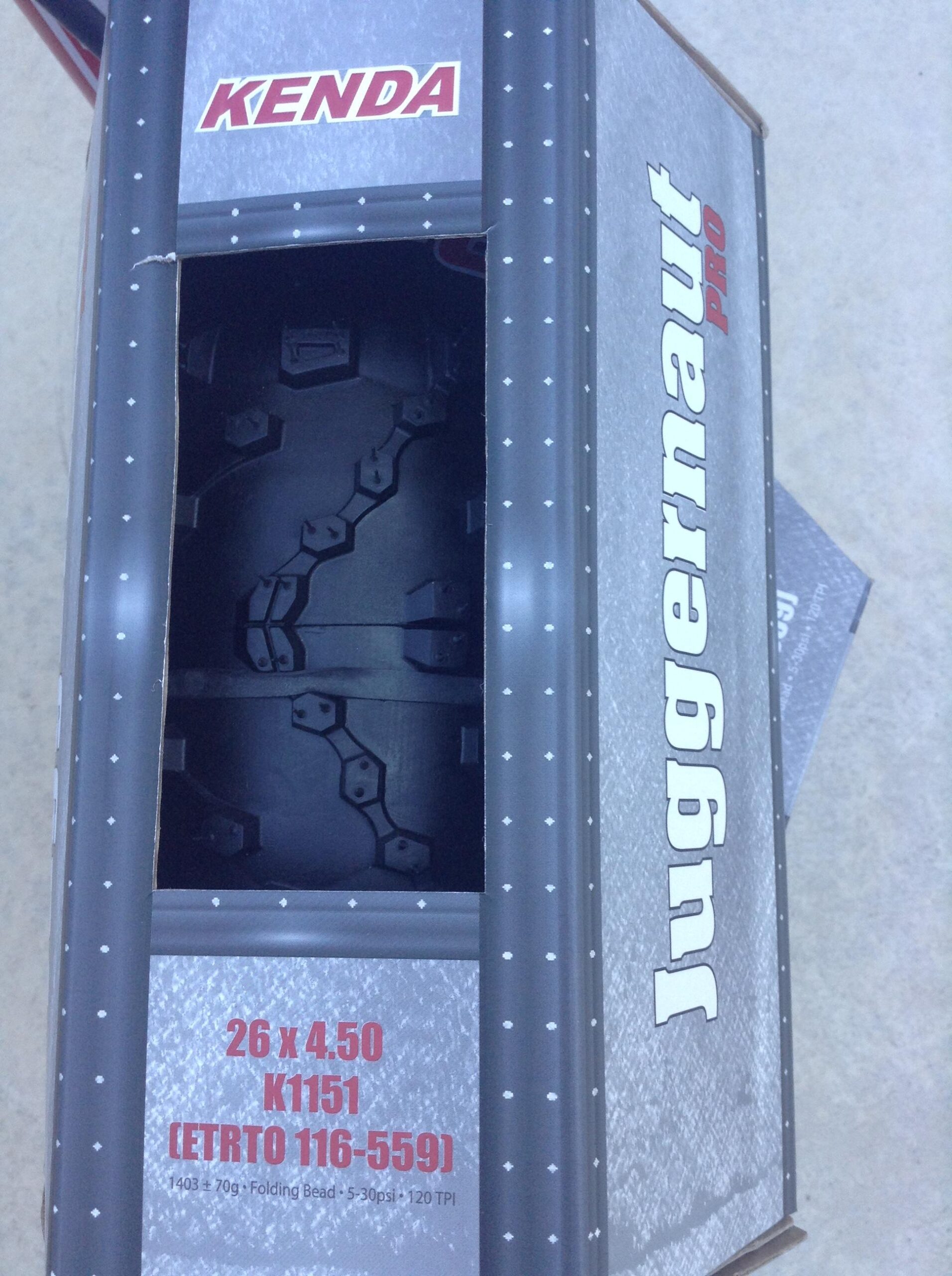 Kenda Juggernaut: Box containing a Kenda Juggernaut Pro tire, featuring a partially open top revealing the tread pattern of the tire inside. The box displays specifications including tire size (26 x 4.50), model number (K1151), and other details in a gray and black design with red accents.