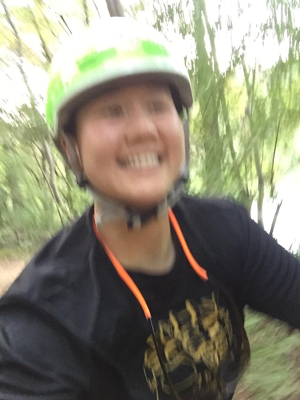A blurred image of a person wearing a green bicycle helmet and a black shirt with a gold design, smiling while riding a bike through a wooded area. Trees and foliage are visible in the background, suggesting an outdoor adventure. USNWC mountain bike trail.