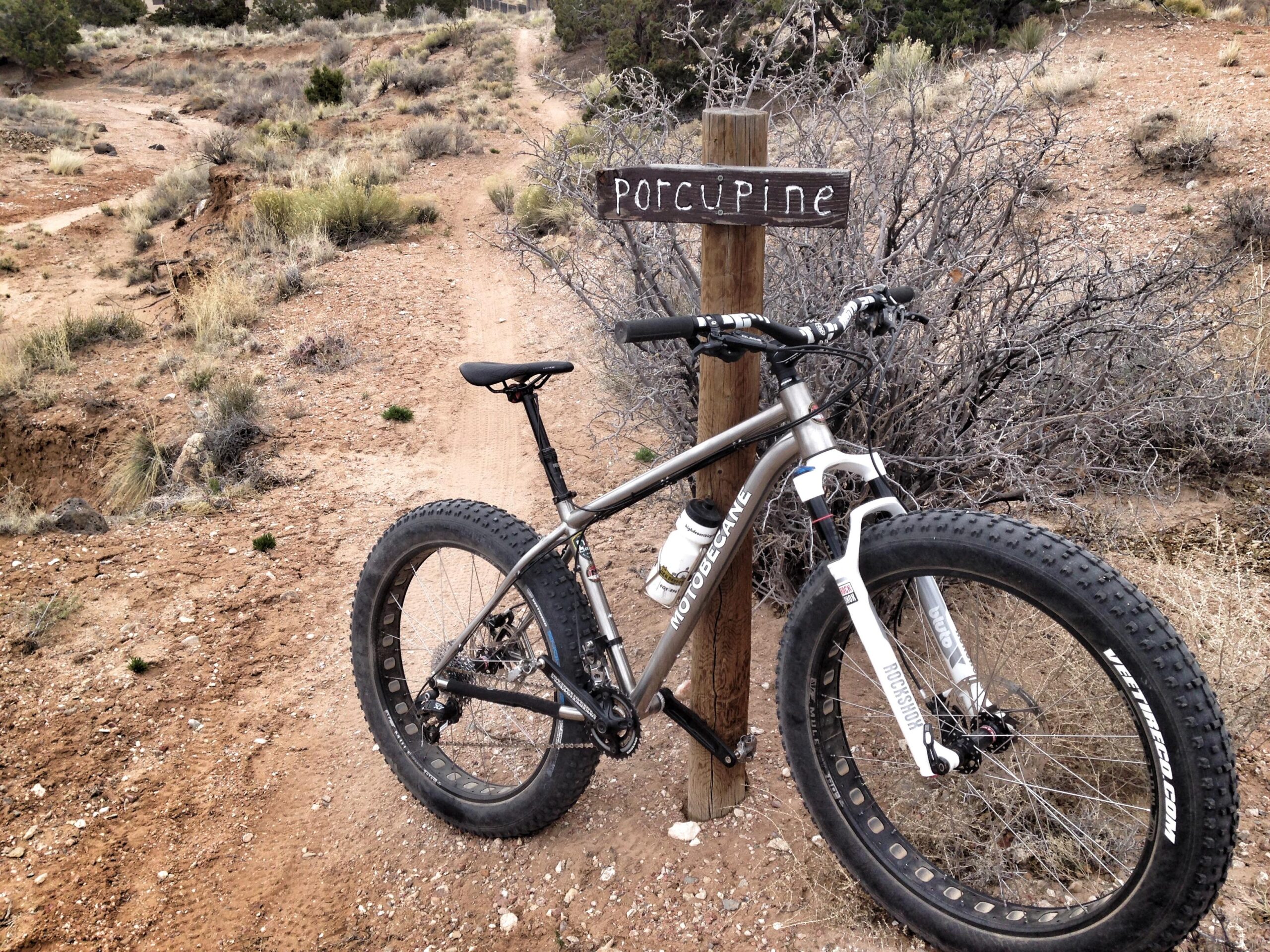 A fat tire mountain bike parked next to a wooden trail sign marked "Porcupine," set against a backdrop of rocky terrain and sparse vegetation. The path leads into the distance, winding through the landscape. Porcupine Fatbike trail mountain bike trail.