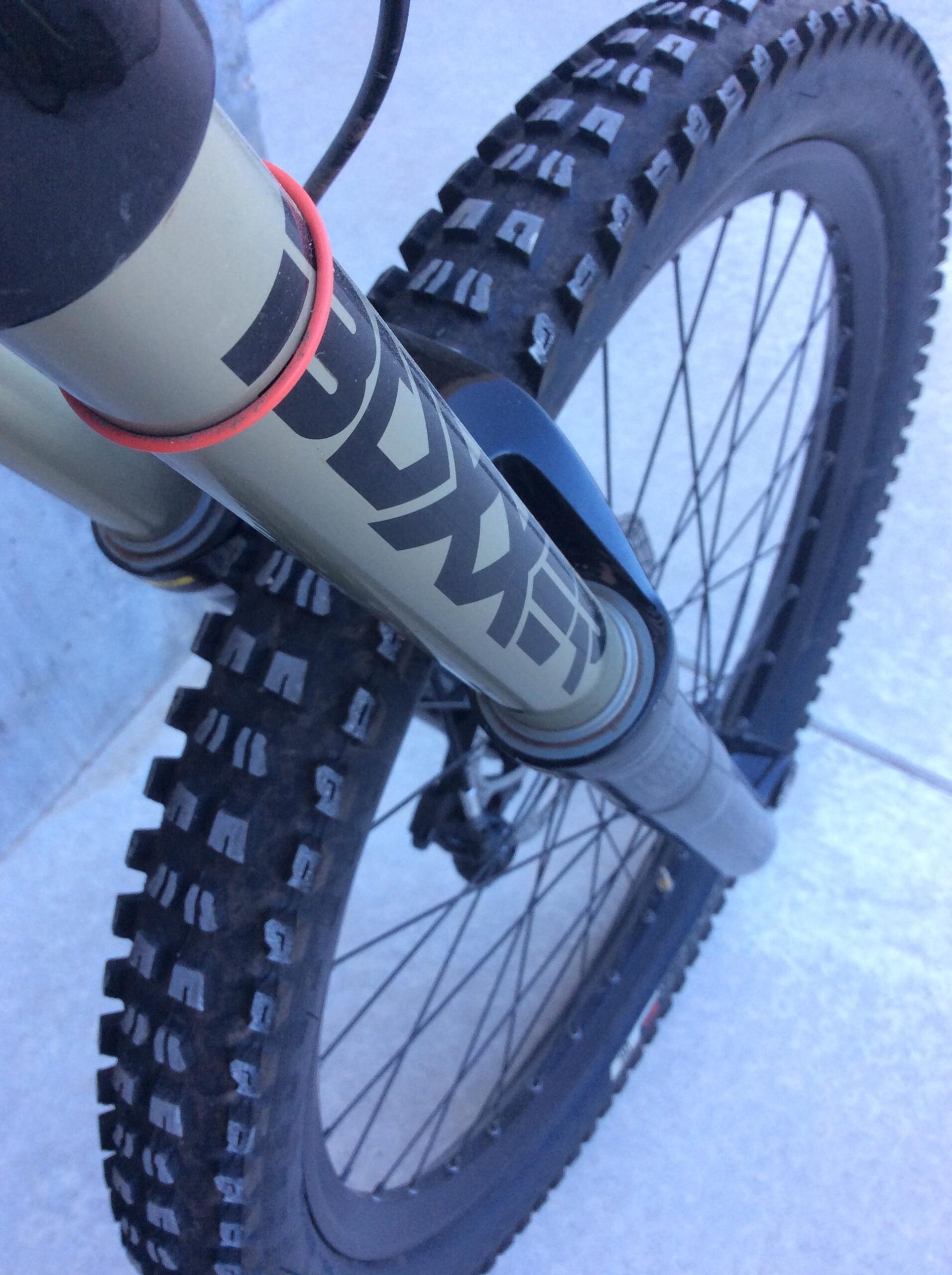 Specialized S-Works Demo 8 Carbon: Close-up of a mountain bike front fork and tire, featuring a green fork with bold graphics, a red rubber band, and a detailed view of a knobby tire designed for traction on rough terrain. The wheel is partially visible, showcasing the tire tread pattern against a light-colored pavement background.