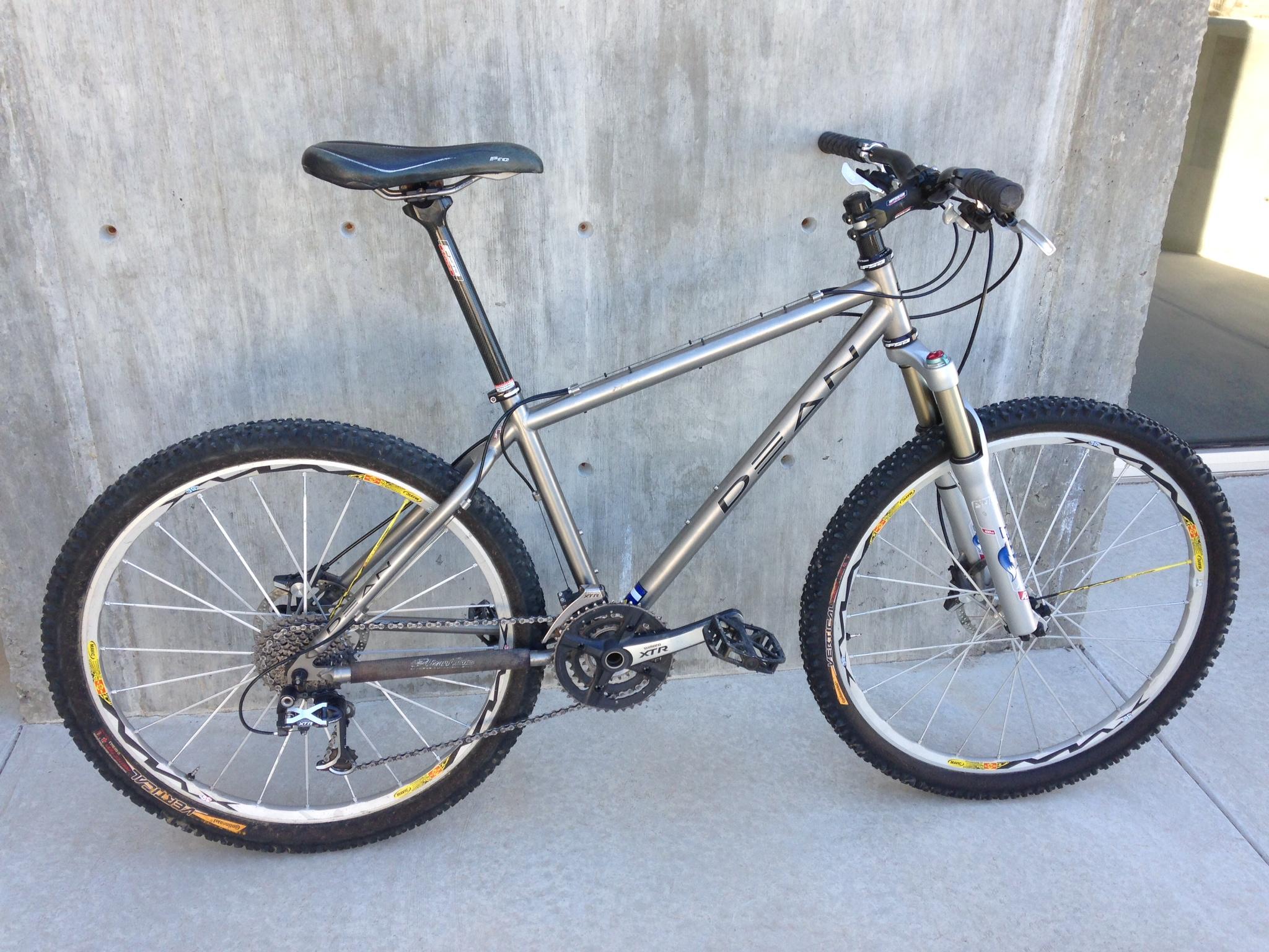 A silver mountain bike with a sturdy frame, featuring thick, textured tires for off-road terrain. The bike has a black seat and handlebars, alongside highlighted components like a front suspension fork. It is positioned against a concrete wall with a smooth surface.