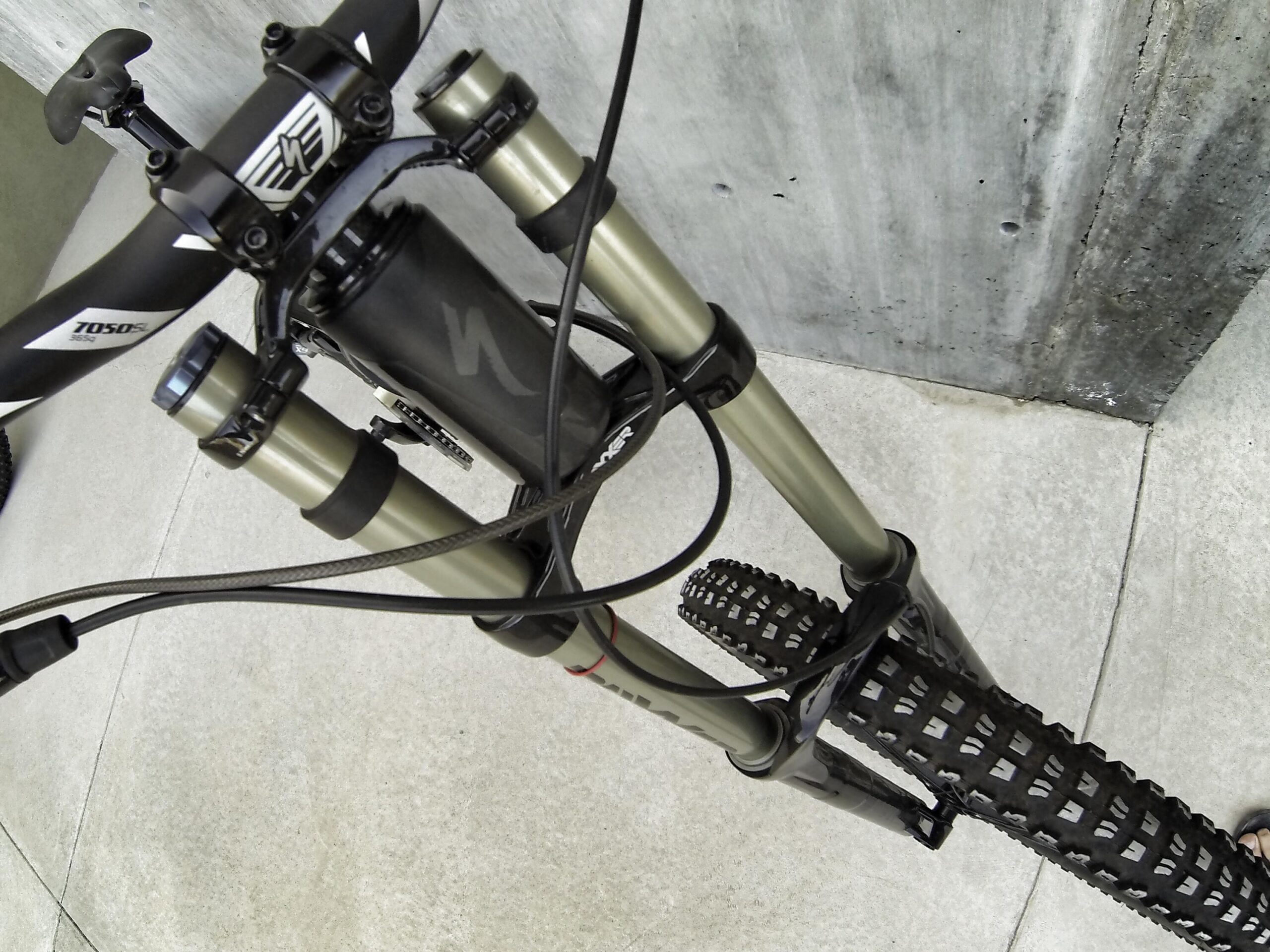 Specialized S-Works Demo 8: Close-up view of a mountain bike's front fork and handlebars, showcasing the suspension system, cables, and tire tread. The background features a textured concrete wall and a light-colored pavement.