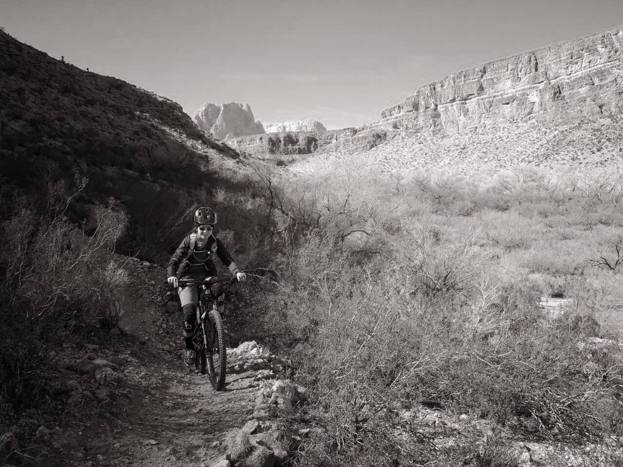 A mountain biker navigating a rocky trail in a desert landscape, surrounded by sparse vegetation and towering cliffs in the background. The image is presented in black and white, emphasizing the rugged terrain and the cyclist's focused expression. Cottonwood Valley North mountain bike trail.