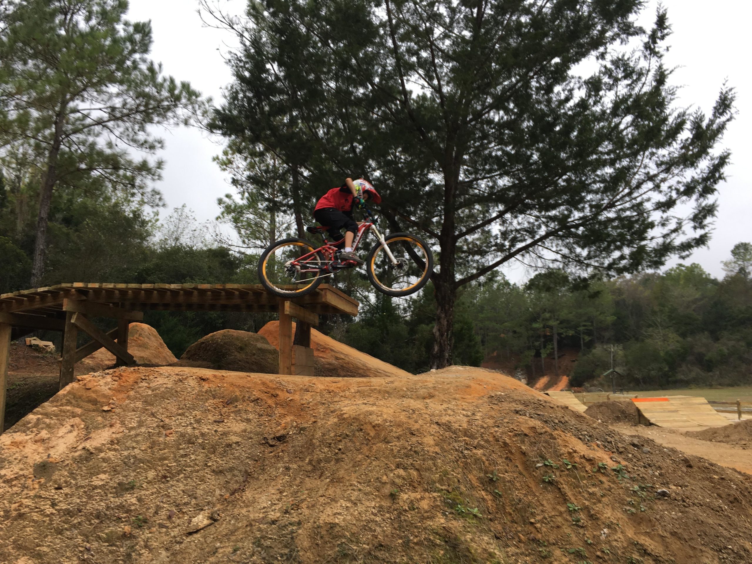 A cyclist wearing a red shirt and helmet performs a jump off a wooden ramp, airborne over a large dirt mound in a bike park surrounded by trees. Santos mountain bike trail.