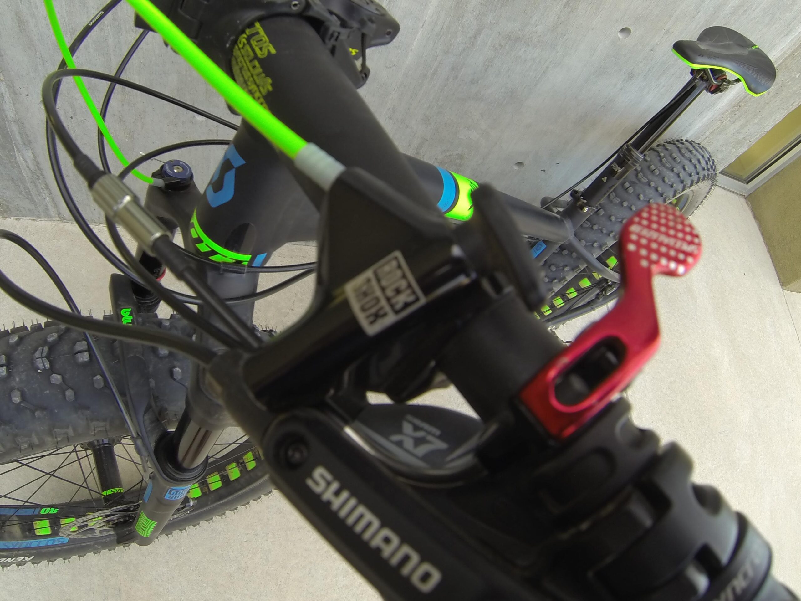 Scott Big Ed: Close-up view of a mountain bike handlebar featuring a Shimano gear shifter and a bright red dropper post lever. The bike's frame is predominantly black with vibrant green and blue accents, and the background shows a concrete wall. The foreground highlights the textured grip and cable systems of the bike, emphasizing its rugged design and functionality.