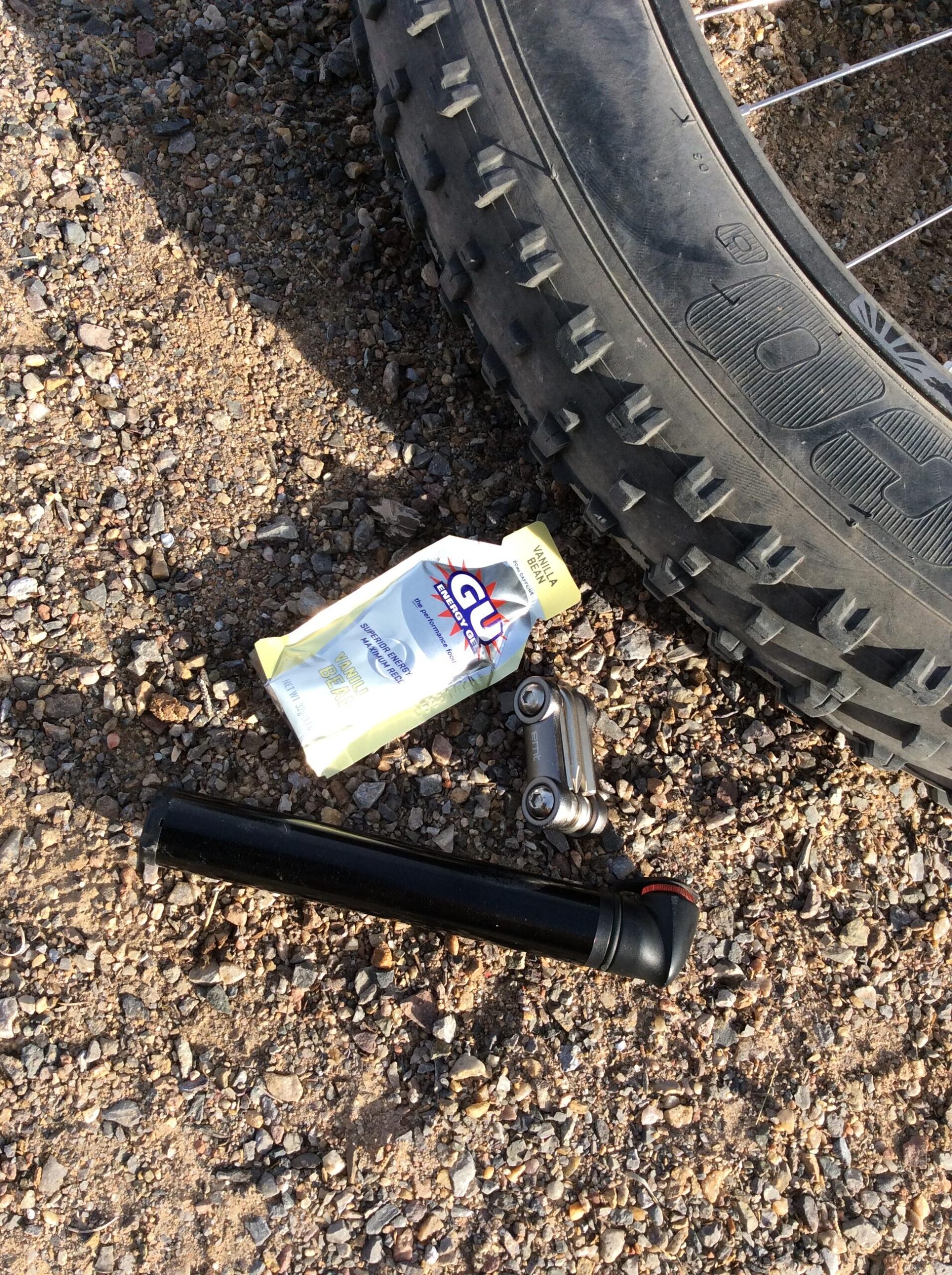 A black bicycle pump, a multi-tool, and a packet of vanilla energy gel, resting on gravel beside a mountain bike tire.