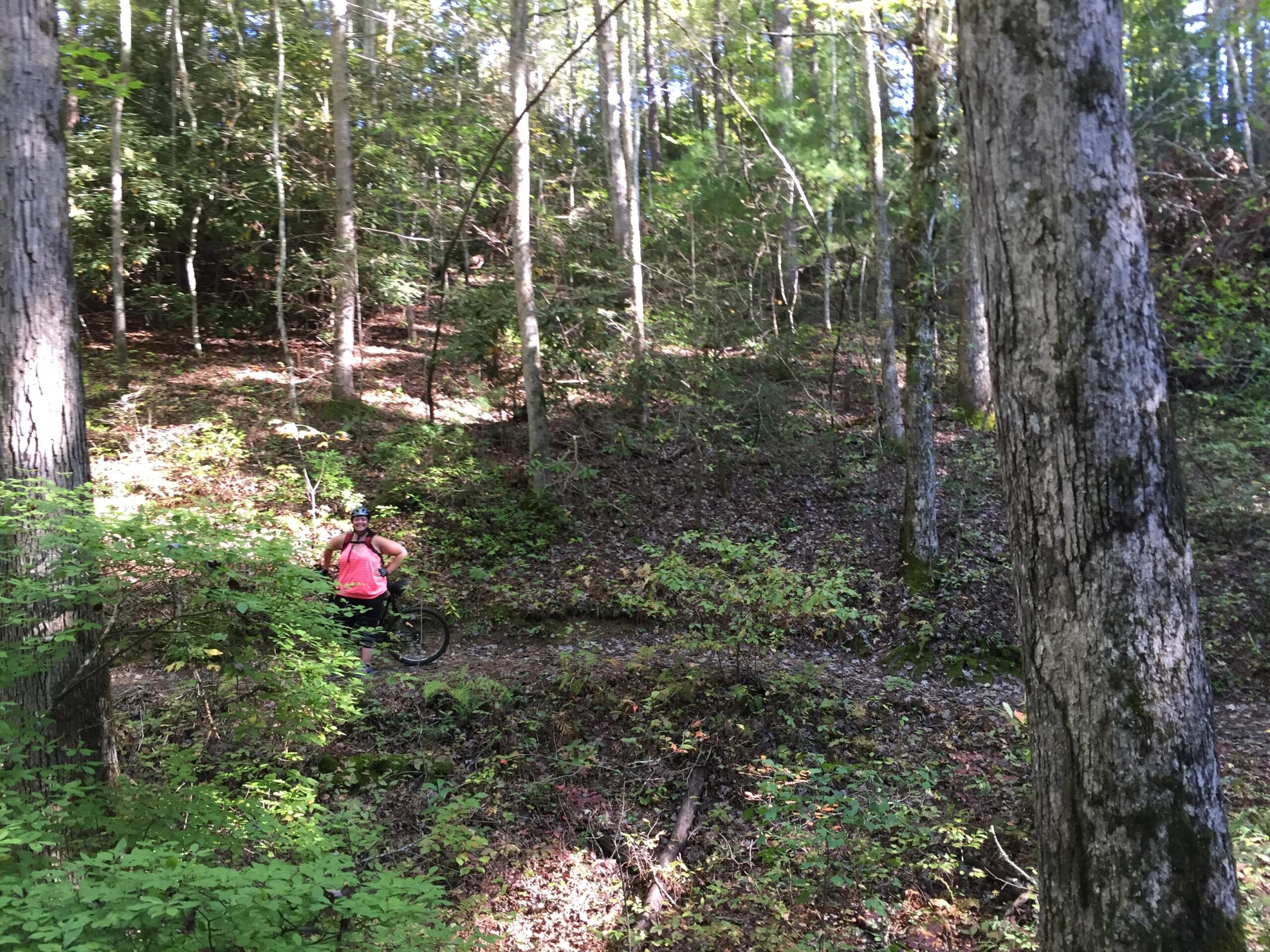 A person wearing a helmet and a pink tank top stands next to a mountain bike on a wooded trail. Surrounding them are tall trees and lush greenery, with sunlight filtering through the leaves, creating a vibrant outdoor scene. Tsali Recreation Area mountain bike trail.