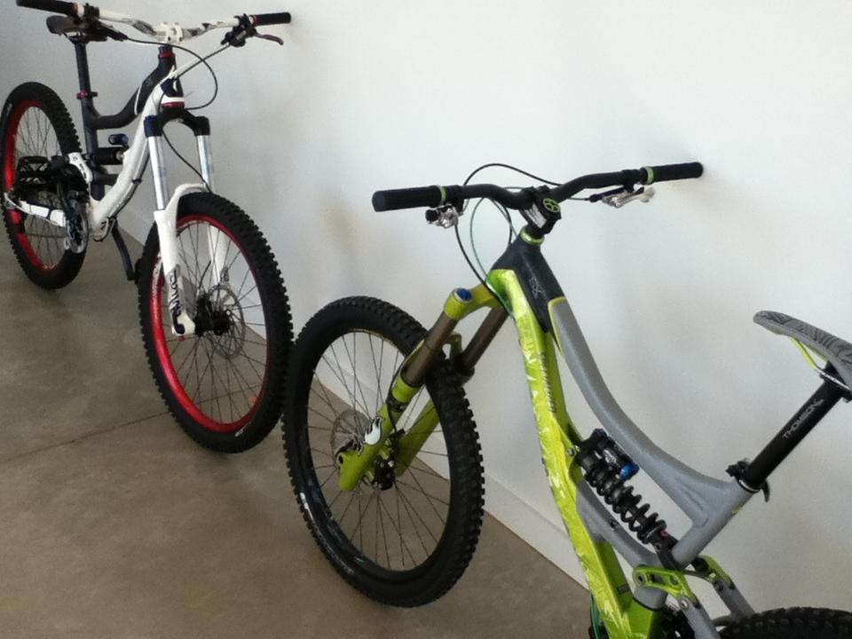 Alt text: Two mountain bikes are positioned side by side against a white wall. The bike on the left features a white frame with red accents and thick tires, while the bike on the right has a green and gray frame, also with thick tires. Both bikes are equipped with handlebars and suspension systems, resting on a concrete floor.