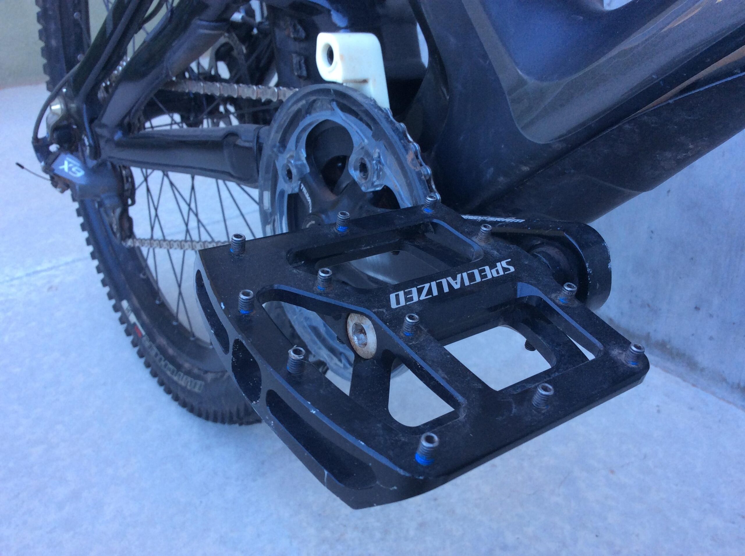 Specialized S-Works Demo 8 Carbon: Close-up of a black bicycle pedal mounted on a mountain bike, showing the pedal's textured surface and the surrounding drivetrain components, including the chain and crankset. The background features a concrete surface, suggesting an outdoor setting.