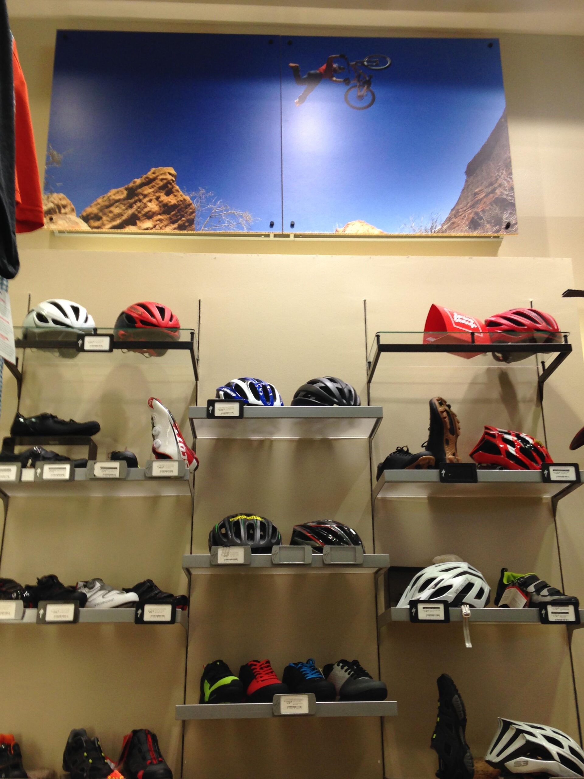 A display of various cycling helmets and shoes on shelves in a store, with a large image of a person performing a bike trick against a blue sky above.