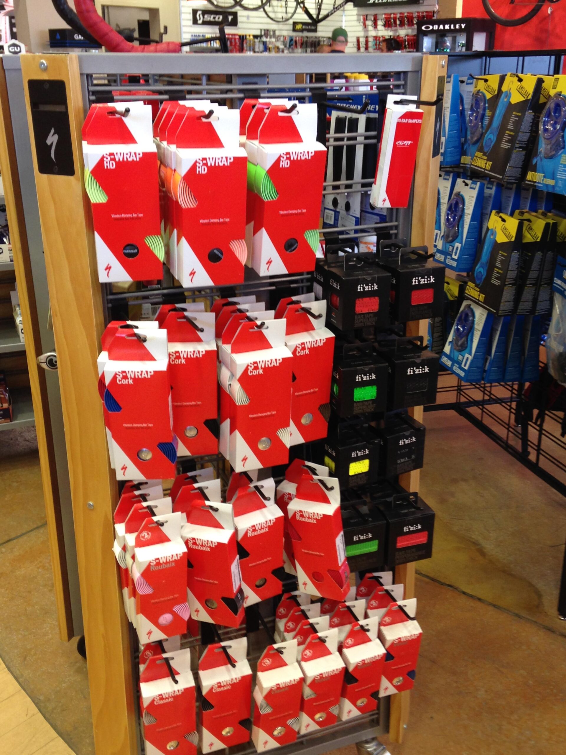 Display of various cycling handlebar tape packages on a retail stand. The packages feature red and white designs, showcasing different colors and types, including "Cork" and "HD" variations. In the background, additional cycling accessories are visible.