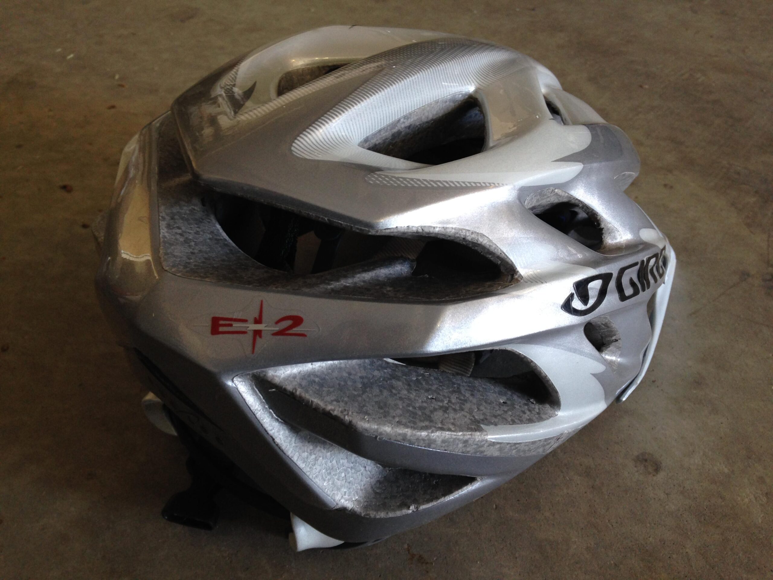 A silver cycling helmet with ventilation holes and a red "E-2" label, resting on a concrete surface.