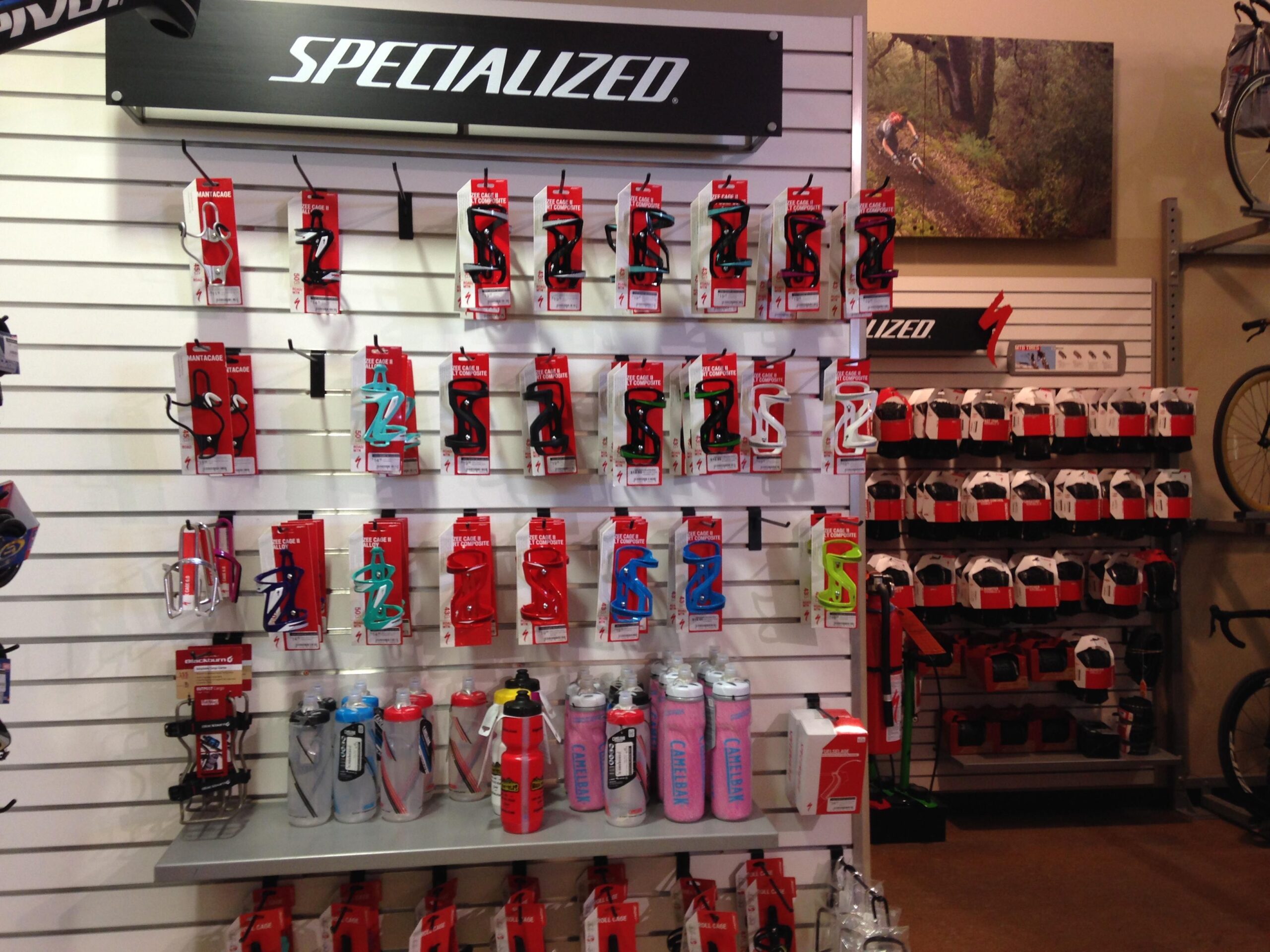 Alt text: A display wall featuring various bicycle water bottle cages and water bottles from the brand Specialized. The items are arranged on hooks and shelves, with red packaging prominently displayed. In the background, there is a photo of a cyclist on a trail and a rack of bike helmets.