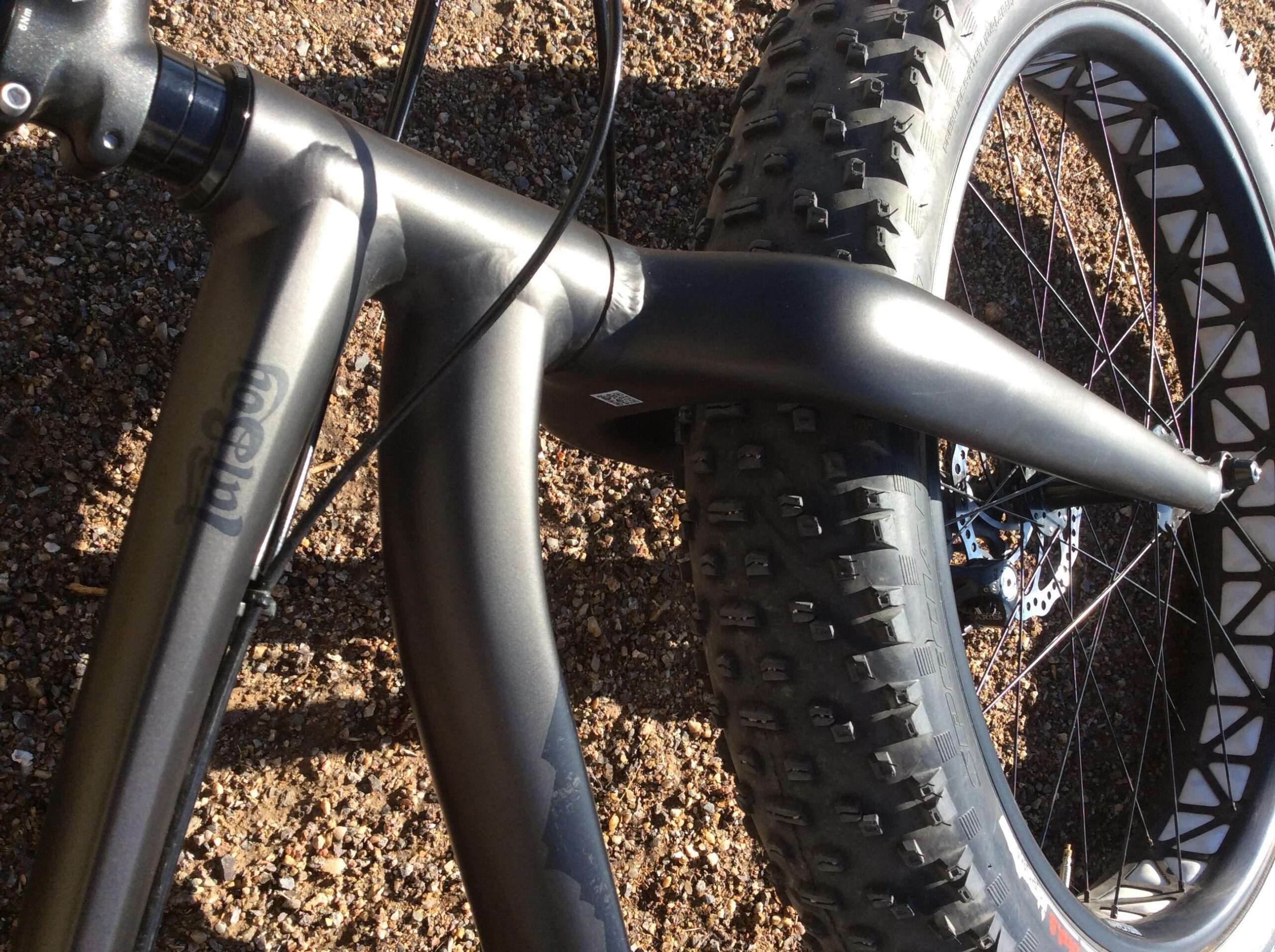 Specialized Fatboy: A close-up view of a fat bike's frame and rear tire, showcasing the matte gray finish of the frame and the textured black tire with visible studs. The background features a gravel surface, enhancing the outdoor cycling context.