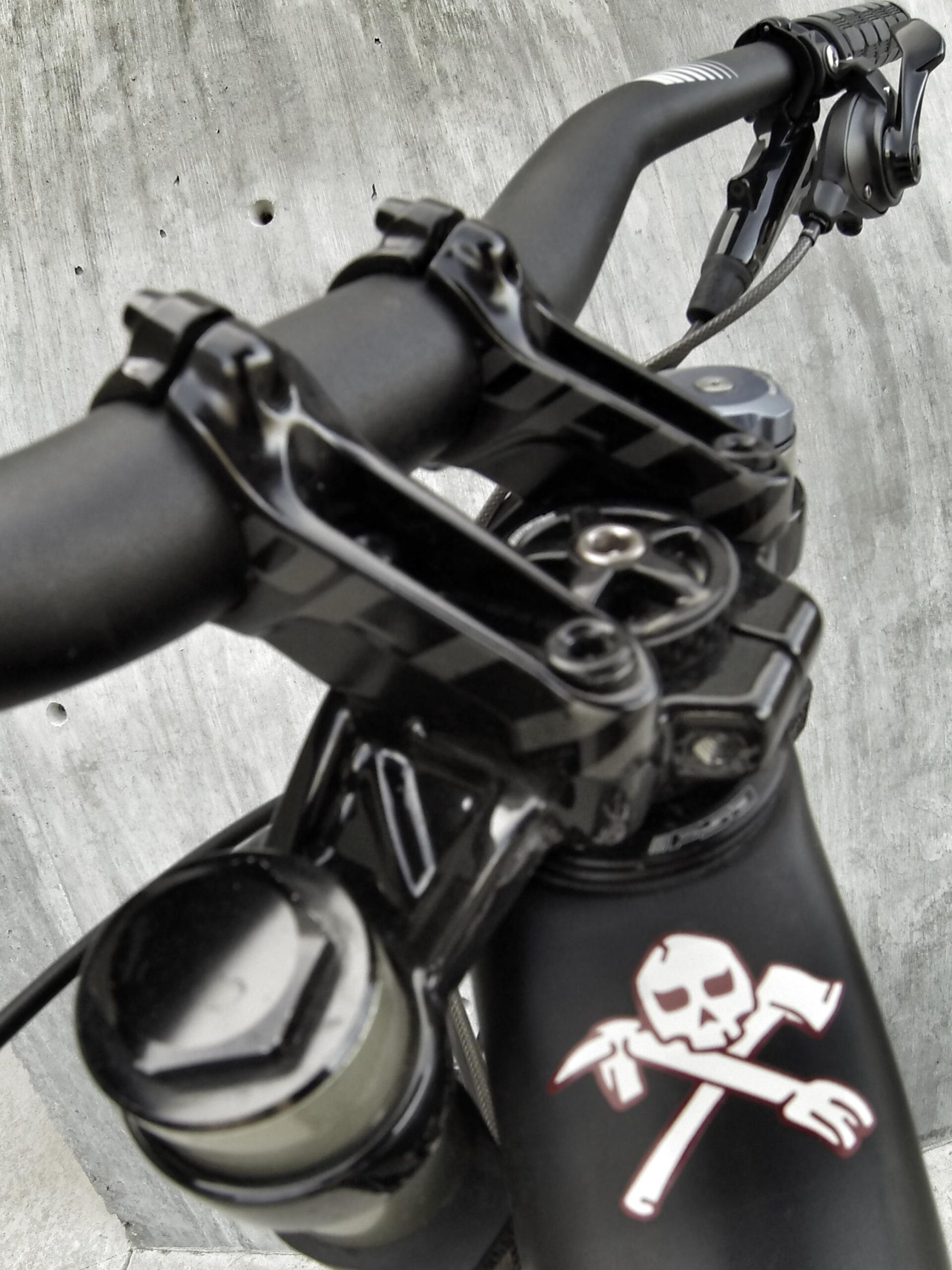 Specialized S-Works Demo 8: Close-up view of a bicycle handlebar featuring a black grip, brake lever, and a distinctive logo with a skull and crossed tools on the frame, set against a textured concrete surface.