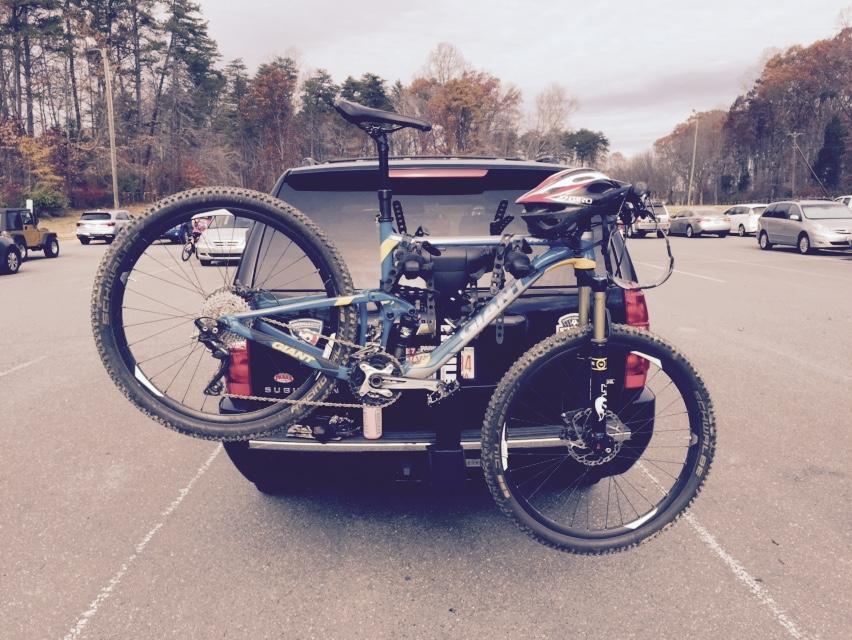Giant Anthem SX 27.5: A mountain bike mounted on a bike rack attached to the back of a vehicle, parked in a lot with several other cars in the background. Trees with autumn foliage are visible in the distance under a cloudy sky.