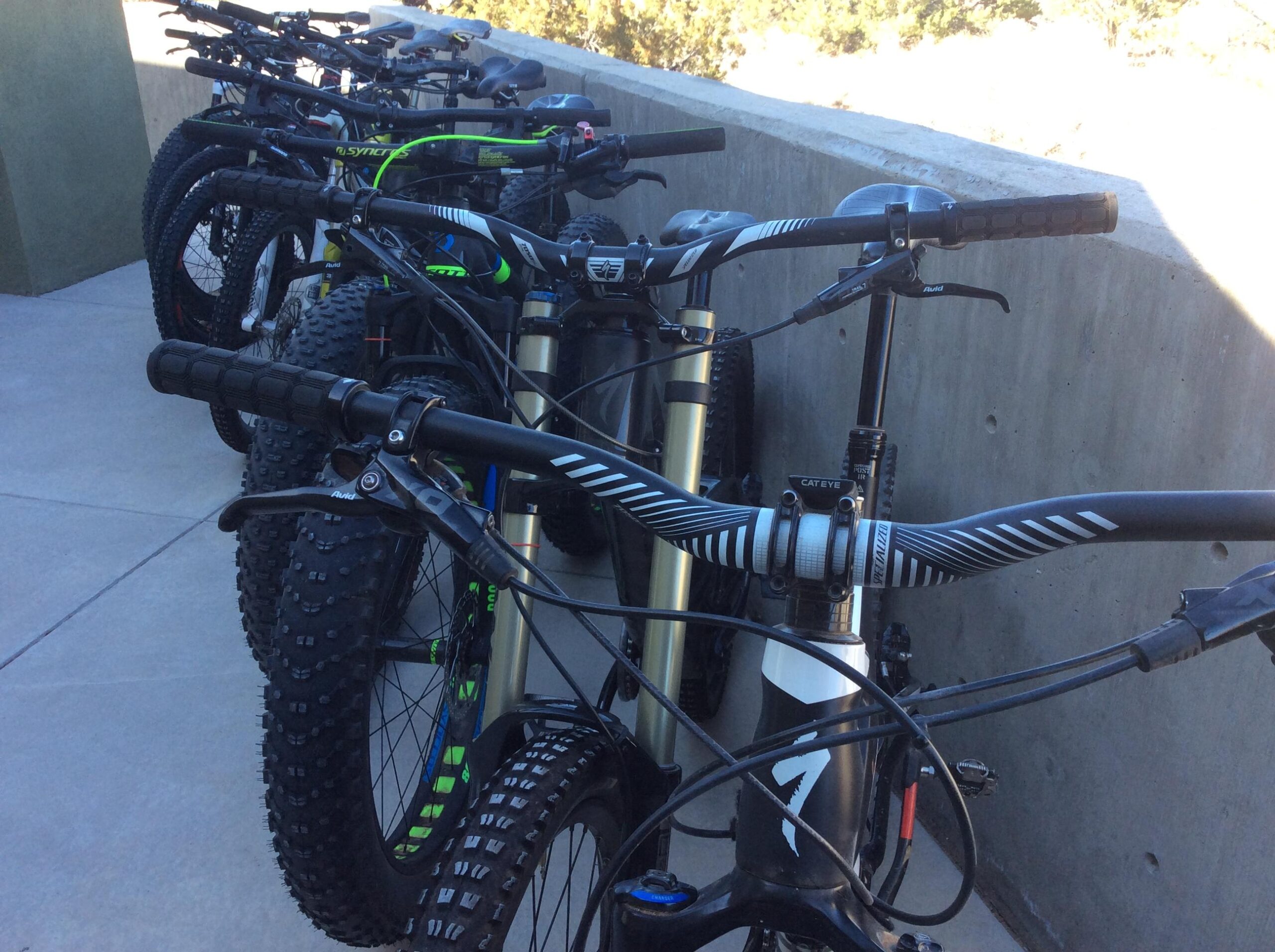 A row of mountain bikes parked on a concrete surface, showcasing their handlebars and tires. The bikes have various frame designs and colors, with some featuring bright accents. The background includes a solid wall and natural greenery.