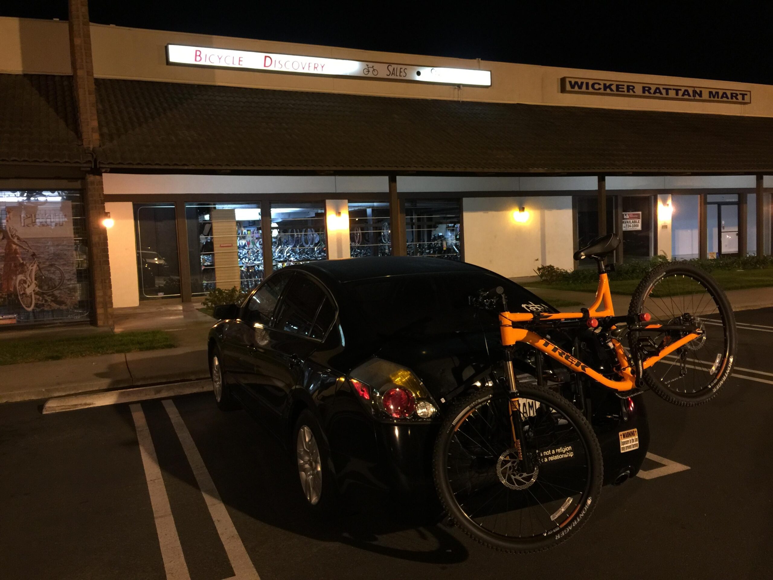 Trek Fuel EX 5 29: A black car parked in front of a bicycle shop at night, with an orange mountain bike mounted on a bike rack at the back. The shop features a sign that reads "Bicycle Discovery," and a window displaying bicycles. Another shop, "Wicker Rattan Mart," is visible in the background.