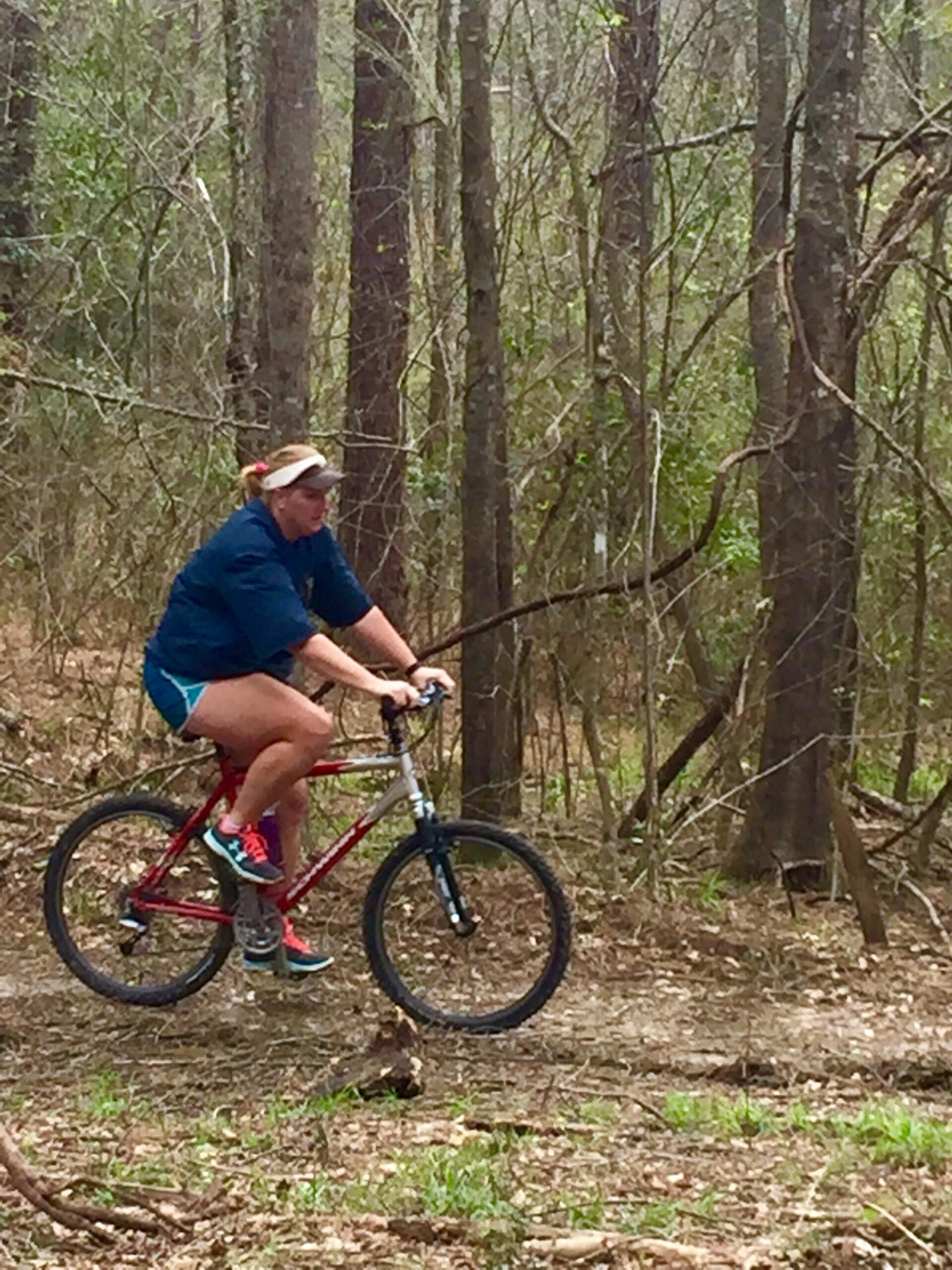 A person riding a mountain bike along a trail in a wooded area, surrounded by trees and natural foliage. The rider is wearing a blue shirt, shorts, and a visor while navigating the uneven terrain. Mt. Zion Bike Trails mountain bike trail.