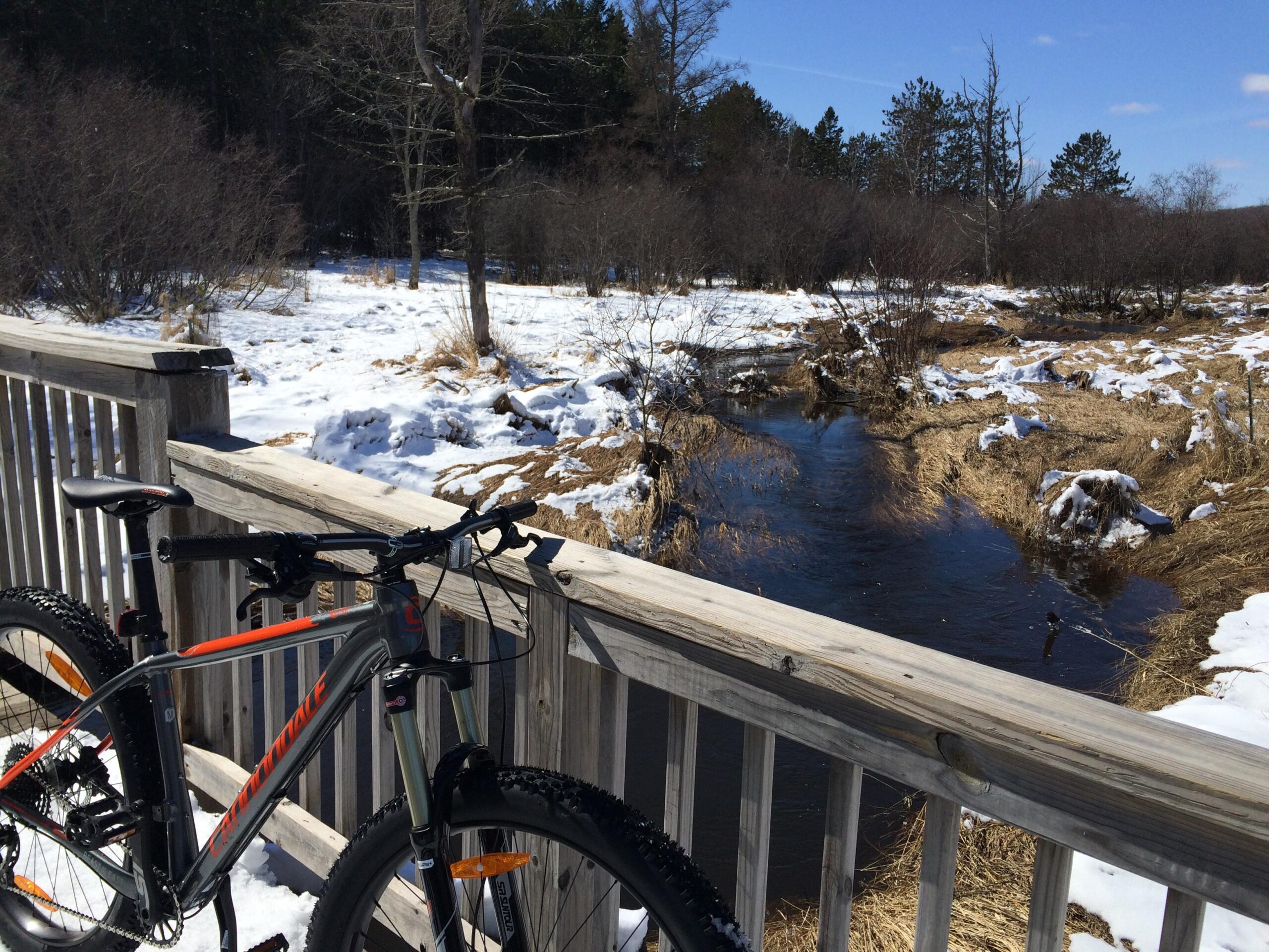 A mountain bike leaning against a wooden railing overlooking a snowy landscape, with a small creek flowing nearby. The scene features patches of snow and dried grass among bare trees, under a clear blue sky. Hartley Park mountain bike trail.