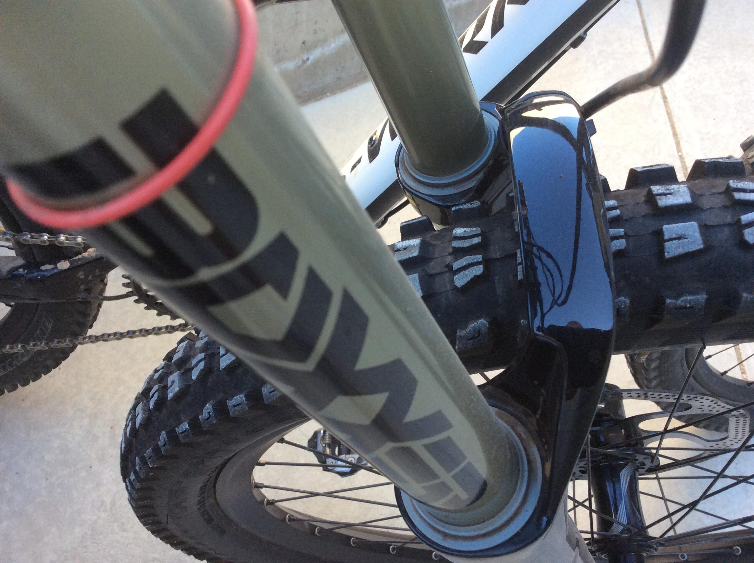 Close-up view of a mountain bike's front suspension fork, showcasing the green fork tubes with markings, an orange rubber ring, and the textured black tire. The bike's frame and chain are partially visible in the background, highlighting the details of the bike's components.