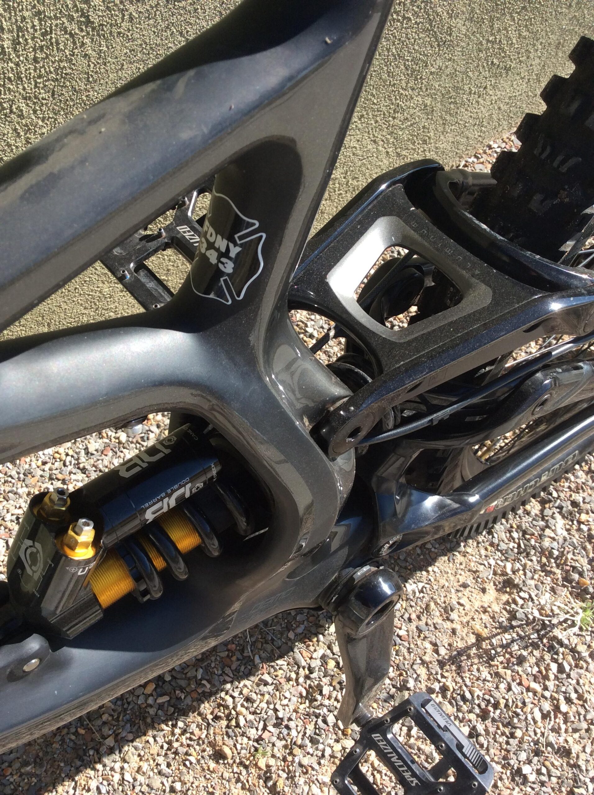 Specialized S-Works Demo 8: Close-up view of a mountain bike frame featuring a suspension system, with a distinctive black and gold shock absorber partially visible. The frame has a matte finish, showcasing a logo and model number on the side. In the background, there is gravel and a textured green surface.