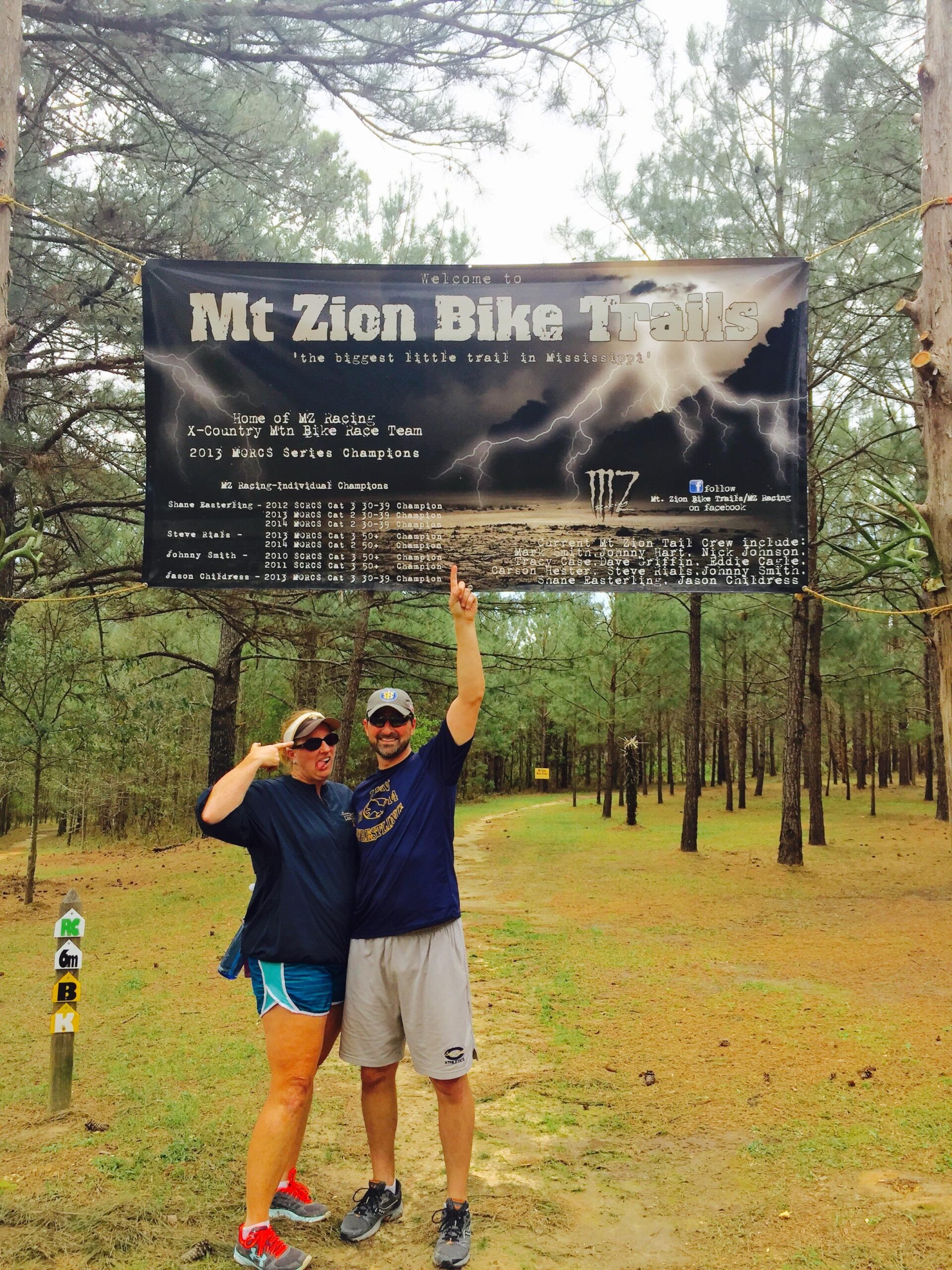 A man and a woman pose playfully in front of a large banner that reads "Welcome to Mt Zion Bike Trails." They are standing on a dirt path surrounded by tall pine trees. The woman makes a silly face while the man raises one hand triumphantly. The path ahead is marked with trail signs. Mt. Zion Bike Trails mountain bike trail.