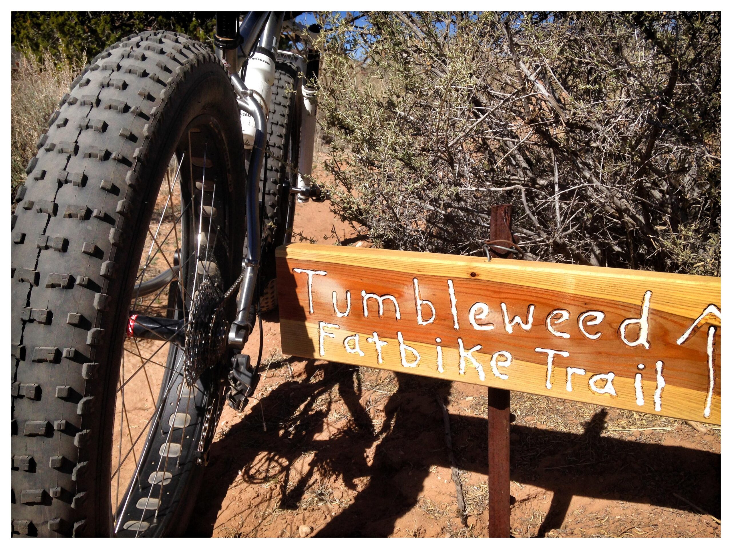 A close-up view of a fatbike tire next to a wooden trail sign that reads "Tumbleweed Fatbike Trail." The sign is positioned on a dirt path surrounded by sparse vegetation, with sunlight casting shadows on the ground. Parkway Fatbike trail mountain bike trail.