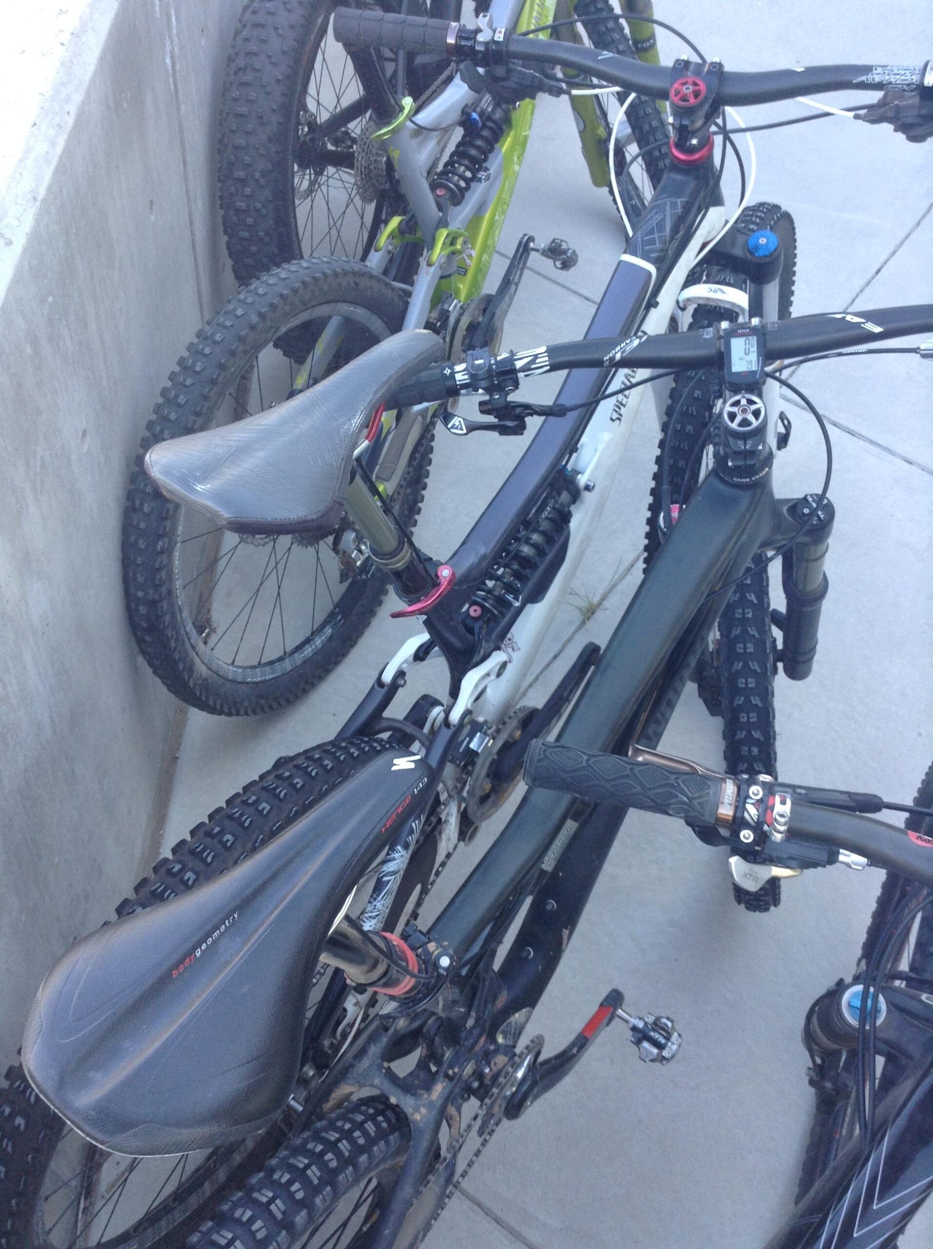 A close-up view of several mountain bikes parked together. The image shows the handlebars, seats, and tires of a few bikes, highlighting their features and details, such as the textured tires and grips. The bikes are leaning against a concrete surface with a slight shadow.