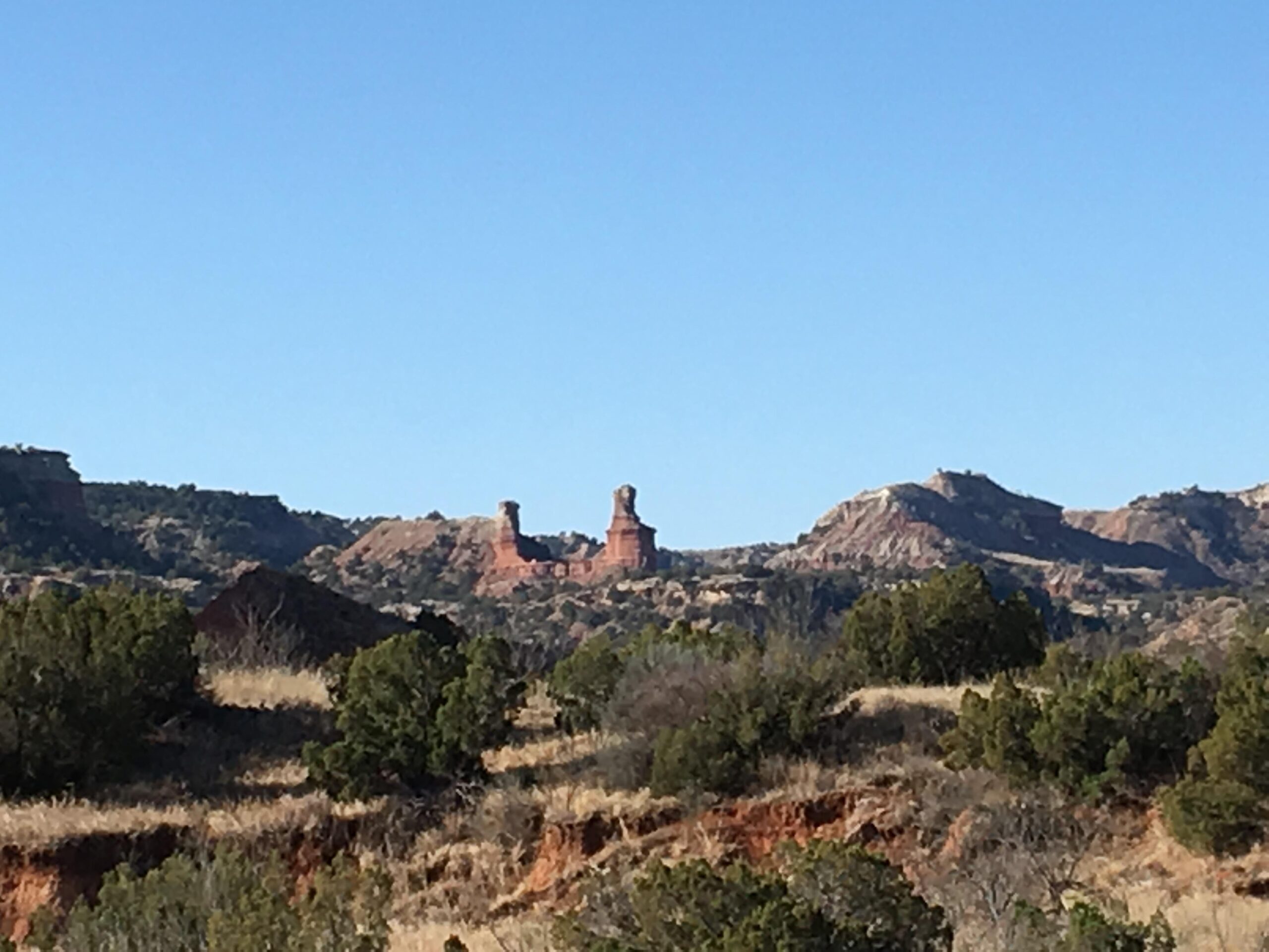 A scenic view of red rock formations set against a clear blue sky, with green shrubs and grass in the foreground and layered hills in the background. The prominent rock structures resemble unique towers in the landscape. Palo Duro Canyon mountain bike trail.