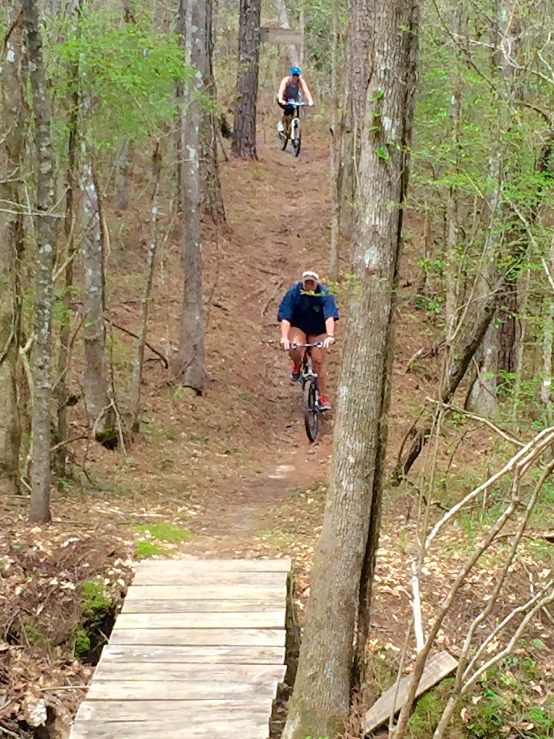 Two mountain bikers navigating a dirt trail in a wooded area. One rider is descending a small hill towards a wooden bridge, while the other is further back on the trail. Surrounding trees are lush with new greenery, indicating springtime. The scene captures the essence of outdoor adventure and nature. Mt. Zion Bike Trails mountain bike trail.