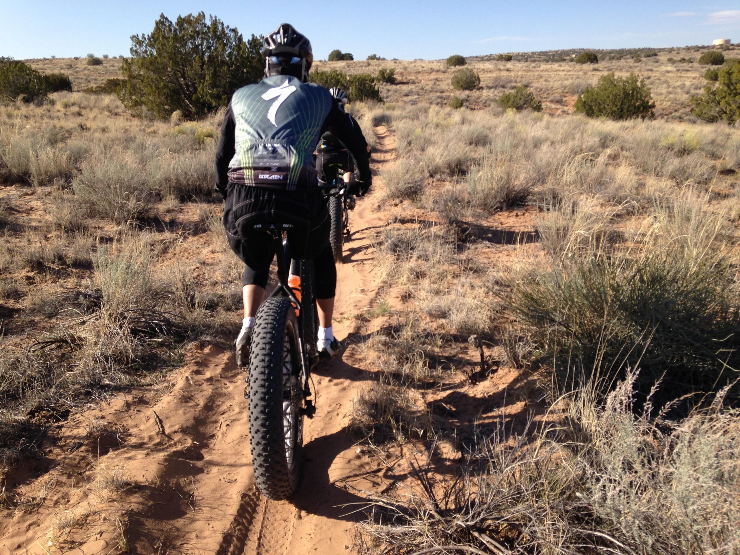 A person riding a fat bike on a sandy trail surrounded by sparse vegetation and shrubs, set in a wide, open landscape under a clear blue sky. The cyclist is seen from behind, wearing a black jersey with the number 7 and black shorts. Super Fat Bike Loop mountain bike trail.