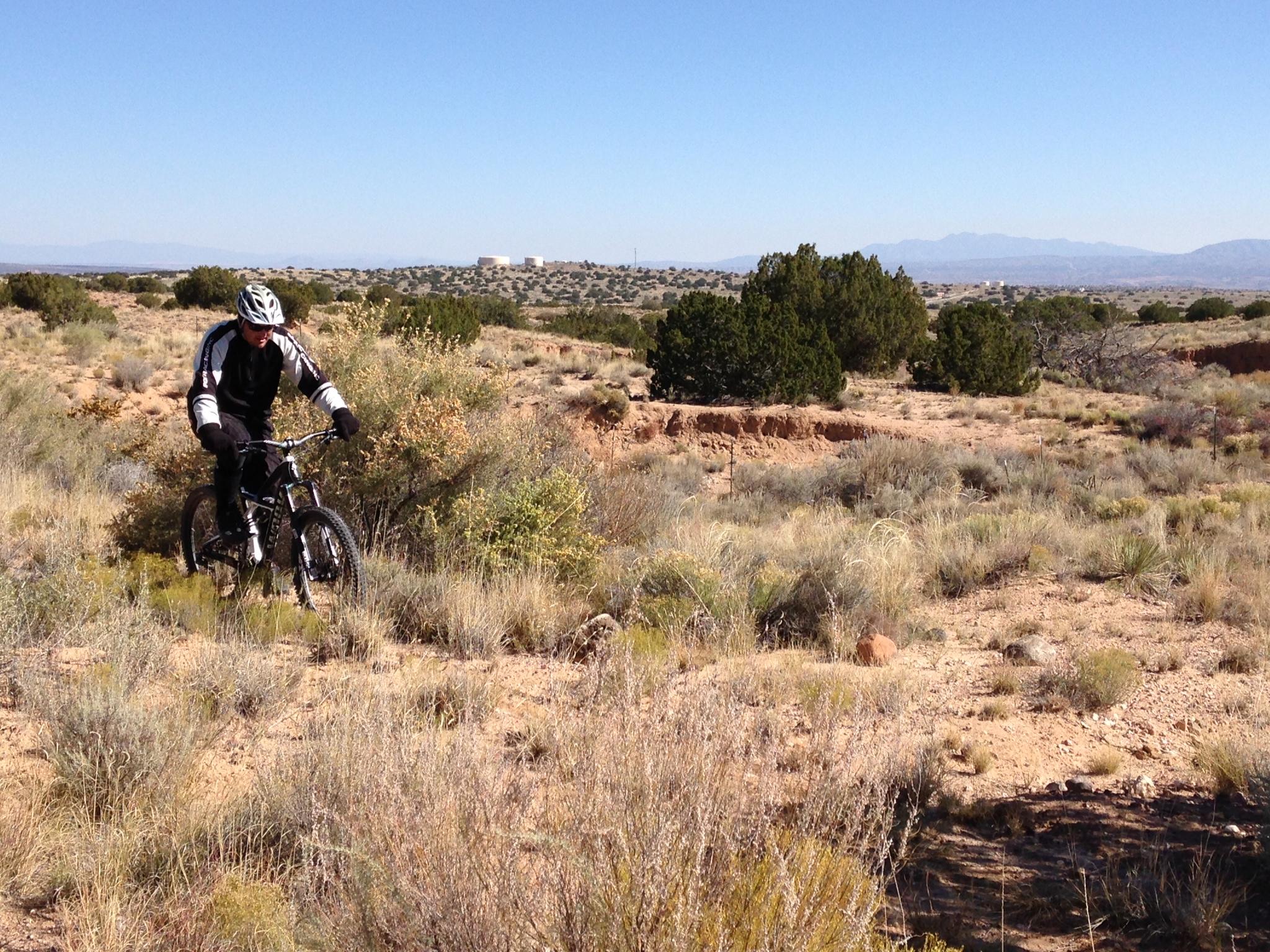 A cyclist dressed in black and white gear rides a mountain bike through a rugged desert landscape, featuring sparse vegetation and distant mountains under a clear blue sky. Parkway Fatbike trail mountain bike trail.